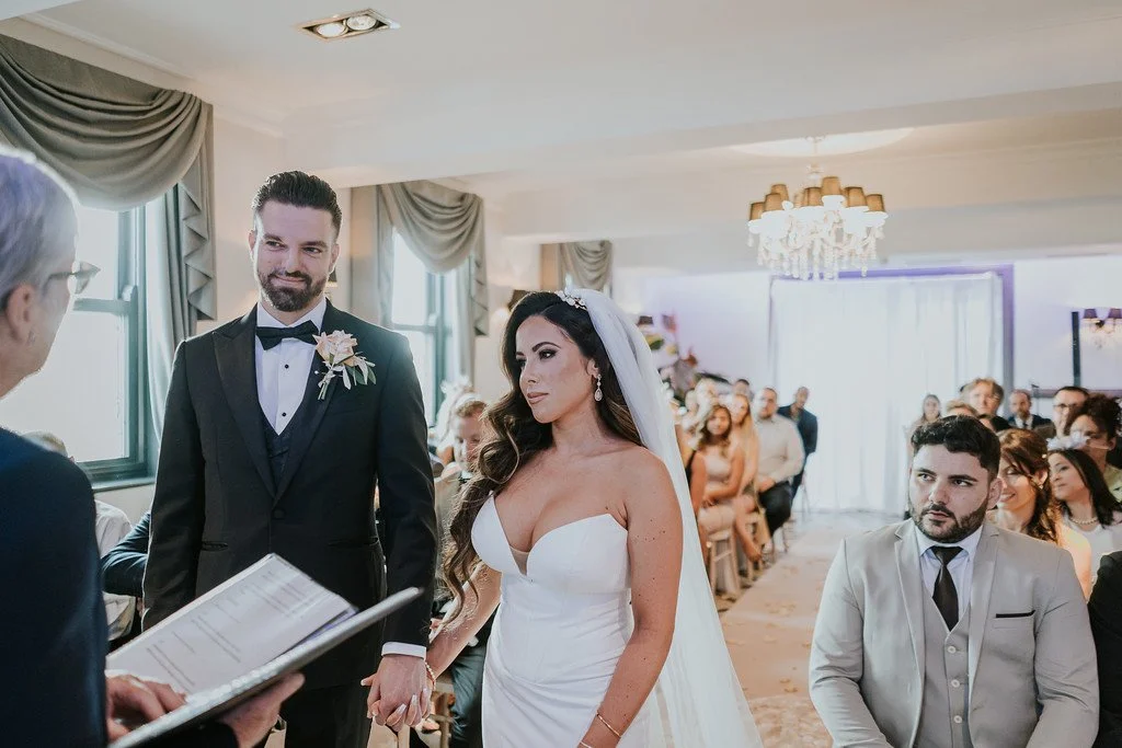 A bride and groom hold hands during their wedding ceremony in a decorated indoor venue, with several guests seated and an officiant presiding.