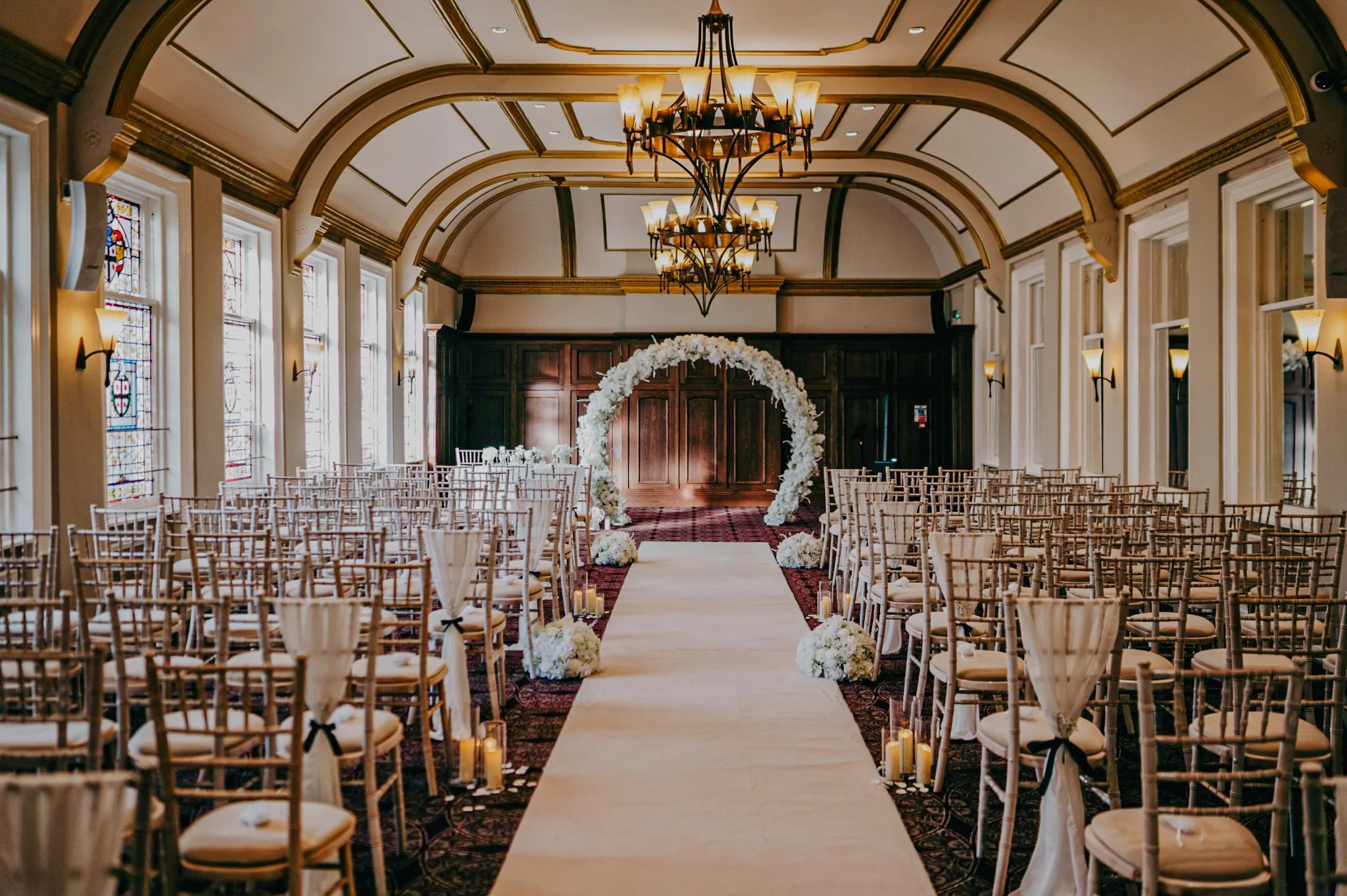 Elegant indoor wedding ceremony setup with rows of chairs decorated with black bows, a floral arch at the altar, warm chandelier lighting, stained glass windows, and candles along the aisle.