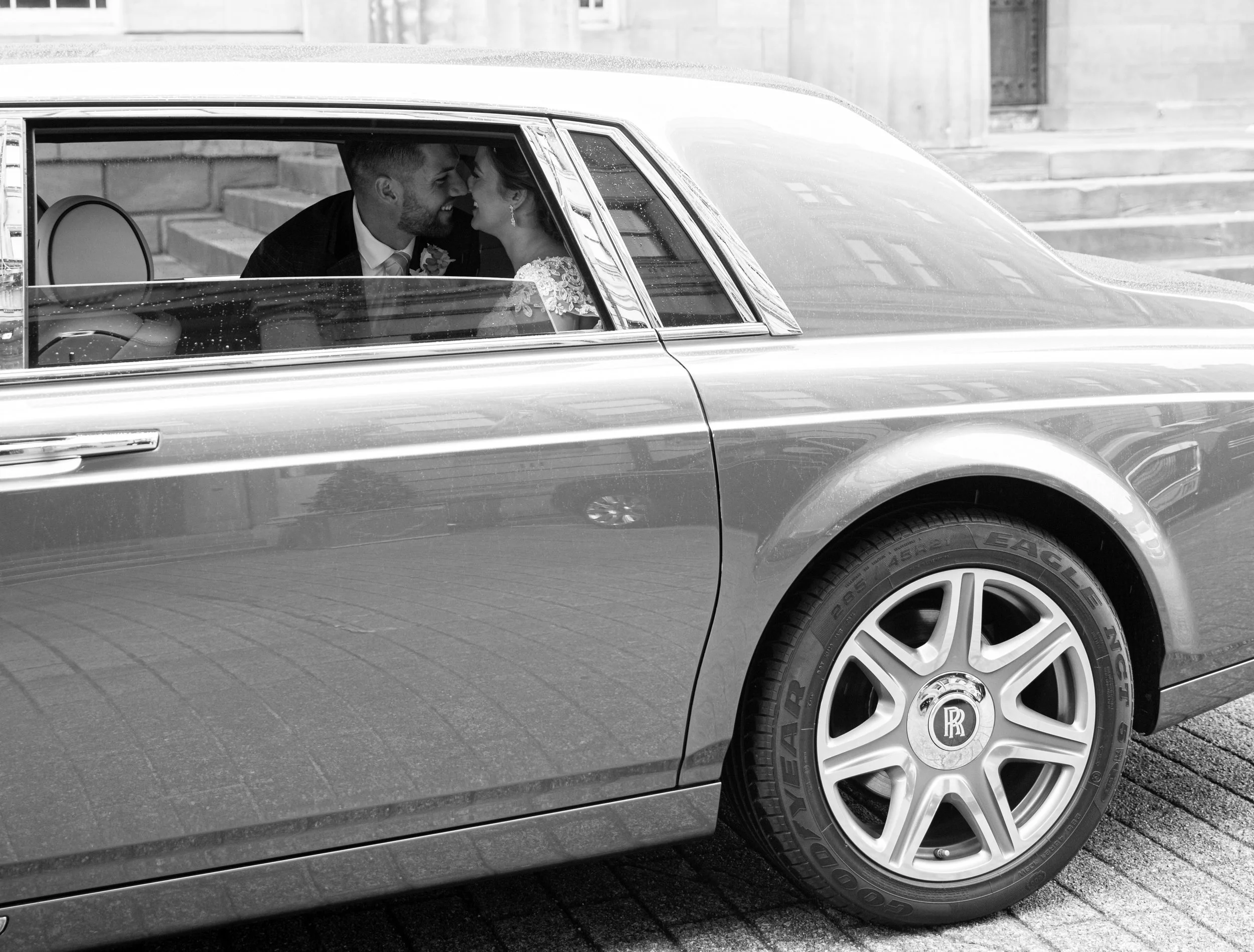 A close-up of a classic luxury car, specifically the front wheel and part of the side. The car is shiny and well-maintained, with visible rain droplets on the surface. Inside the car, a smiling couple dressed in wedding attire are leaning in close to