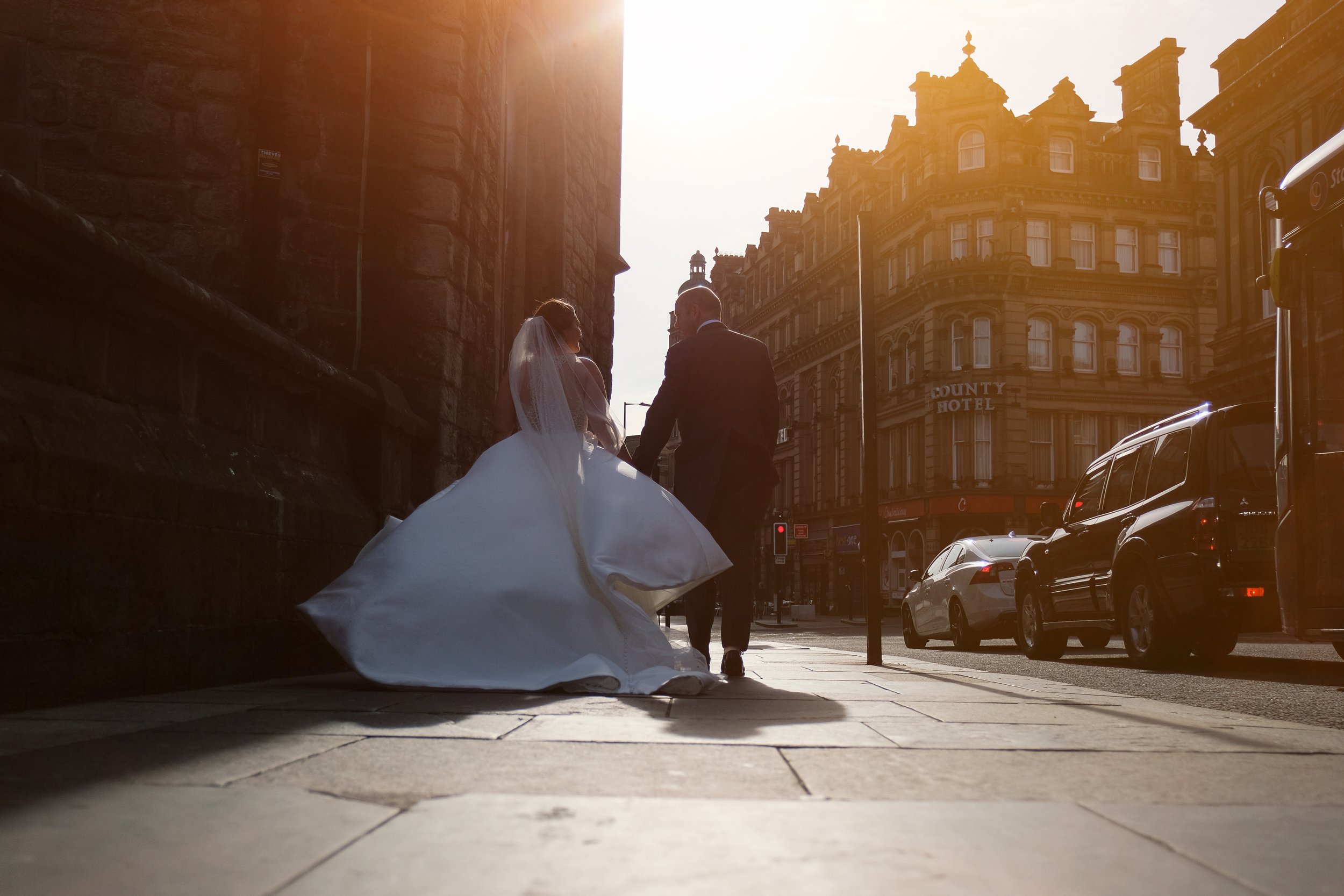 A bride in a gown and veil holding hands with a groom in a suit walking on a city sidewalk during sunset, with historic building facades and parked cars in the background.