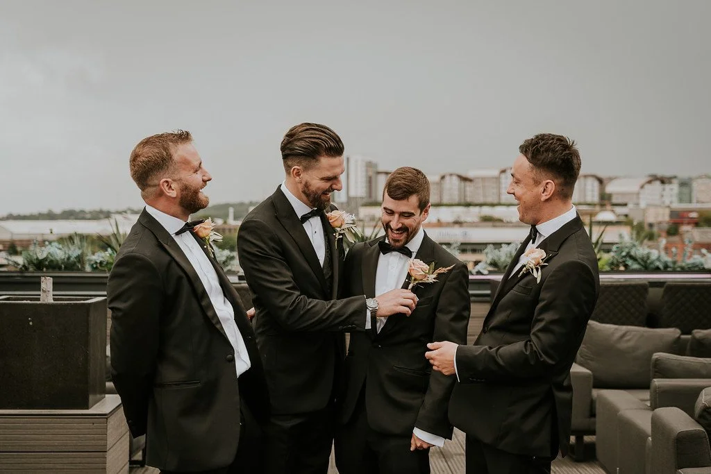 Four men in tuxedos enjoying a lighthearted moment on a rooftop with city buildings in the background.
