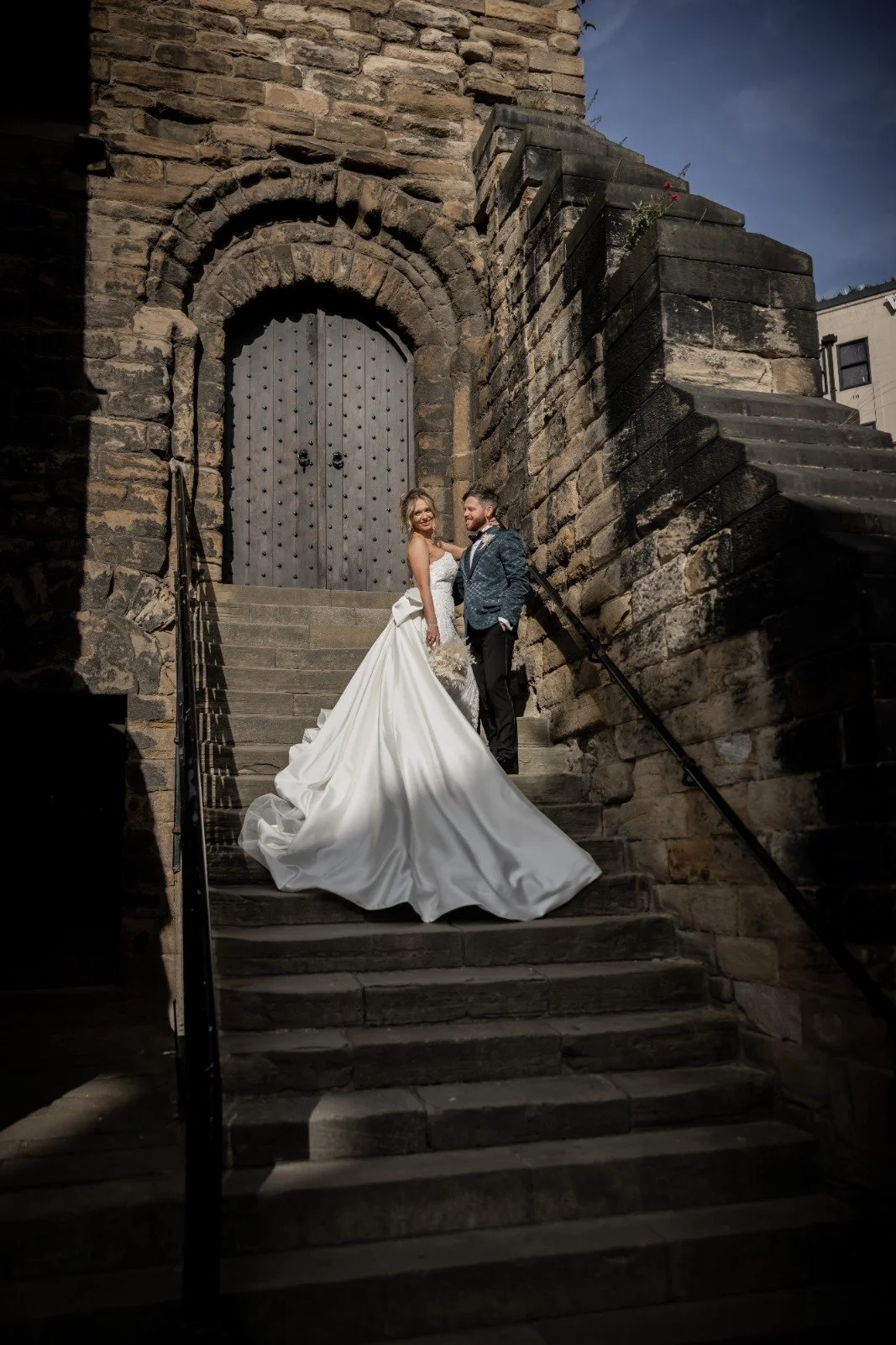 A bride and groom on stone steps in front of a large wooden door, with a stone wall arching above. The bride is wearing a white wedding gown with a long train and holding a bouquet. The groom is dressed in a patterned suit jacket and black trousers. 