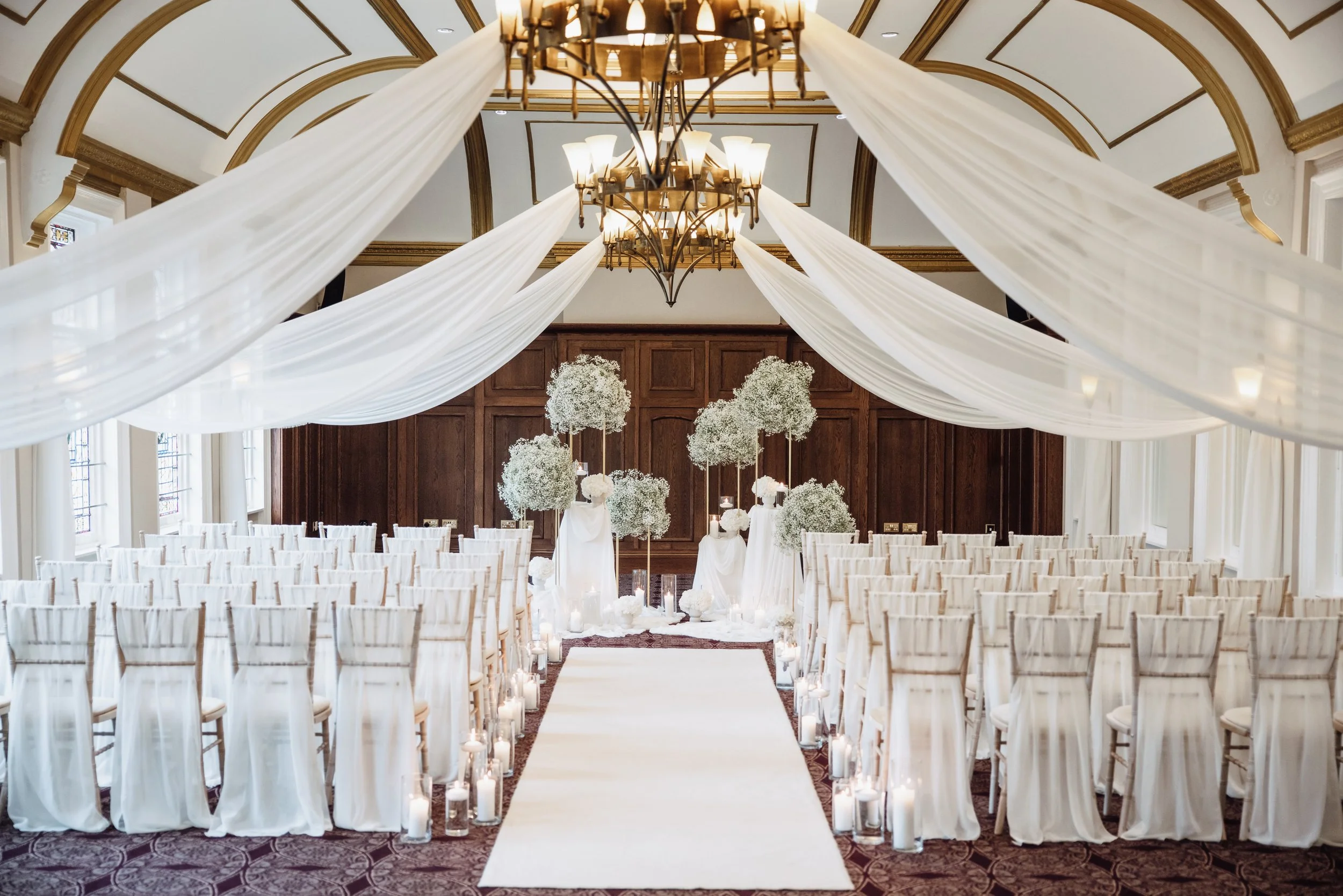 Elegant wedding ceremony setup with white drapes hanging from the ceiling, floral arrangements with white flowers, seating for guests, a white aisle runner, and candles lining the aisle, all in a grand hall with wooden paneling and high arched ceilin