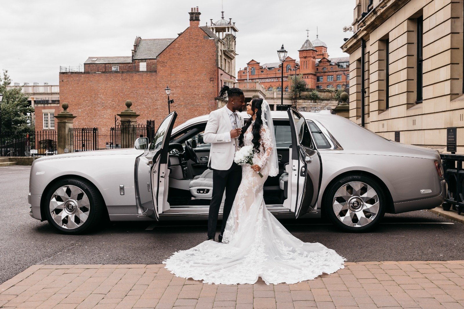 A bride and groom in wedding attire standing in front of a silver luxury car with the doors open, sharing a romantic moment outdoors in an urban setting.