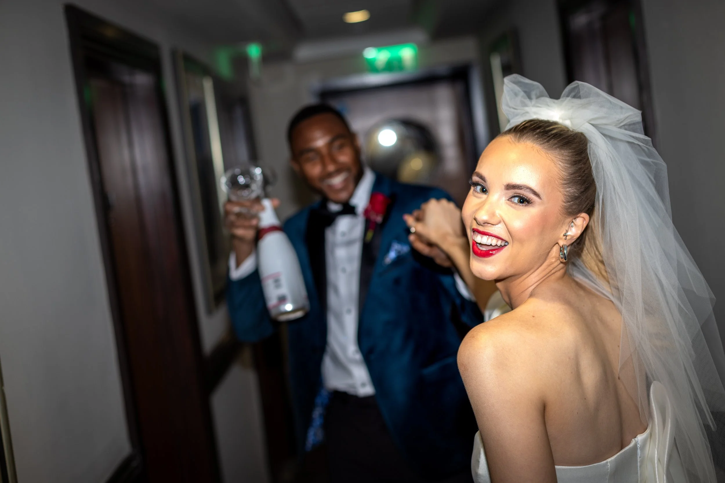 A smiling bride with red lipstick and a veil, posing for a photo in an elevator while a man dressed in a tuxedo in the background points a gun at the camera.