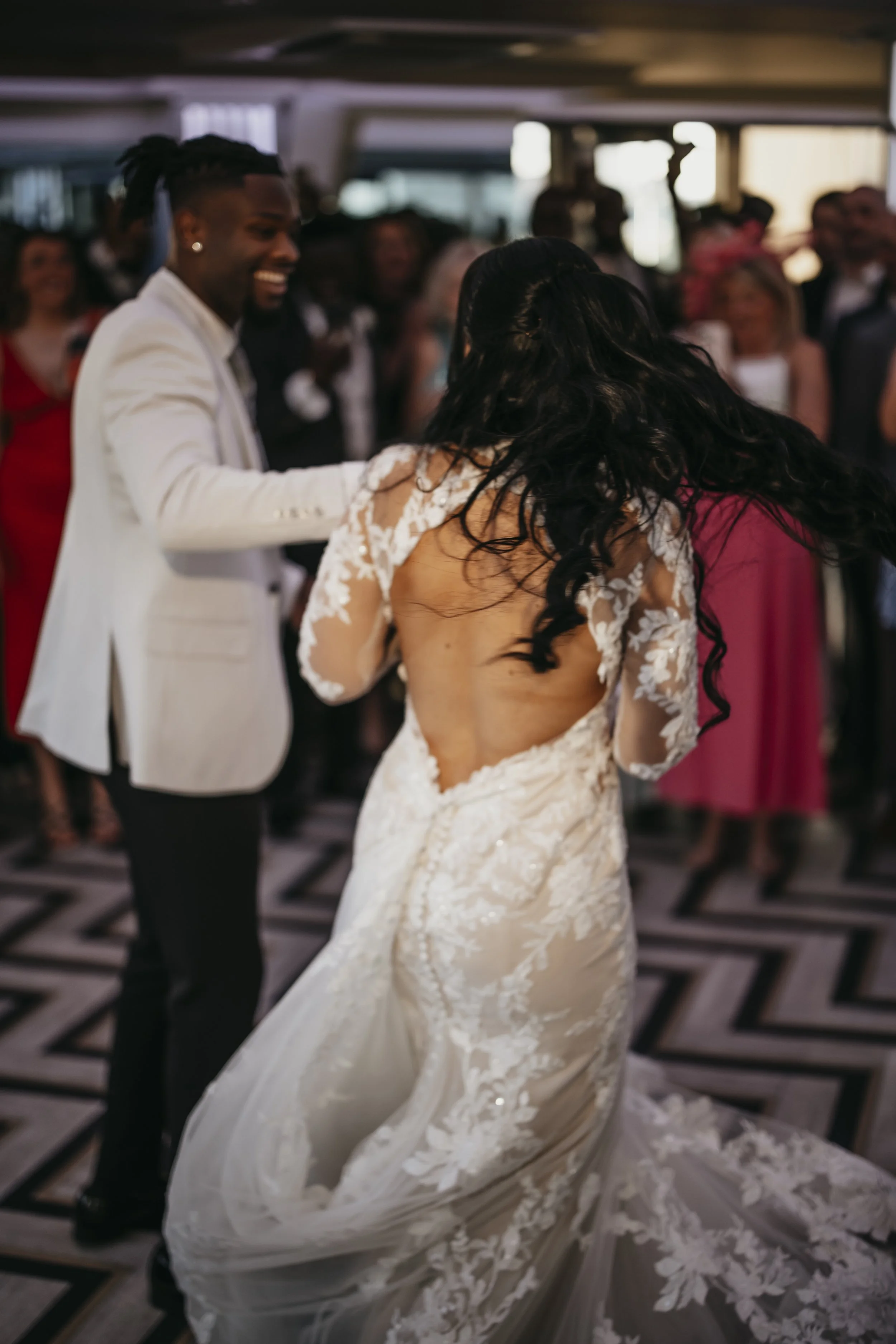 A bride and groom are dancing at their wedding reception. The bride is wearing a lace wedding gown with a long train, and the groom is dressed in a white jacket and black pants. They are surrounded by guests in a decorated indoor venue.