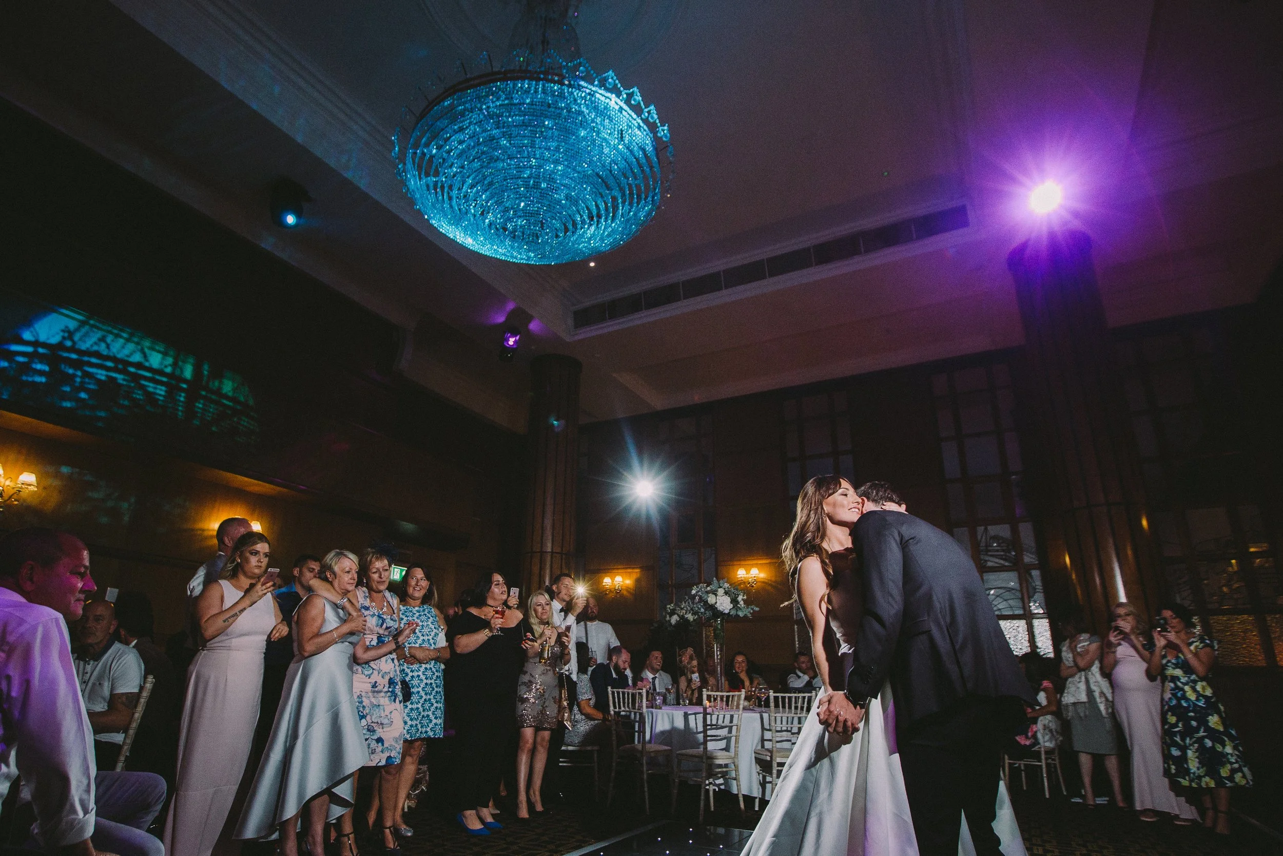 A wedding ceremony with the bride and groom dancing closely in a dimly lit banquet hall filled with guests, a blue chandelier hanging from the ceiling, and colorful lighting effects.