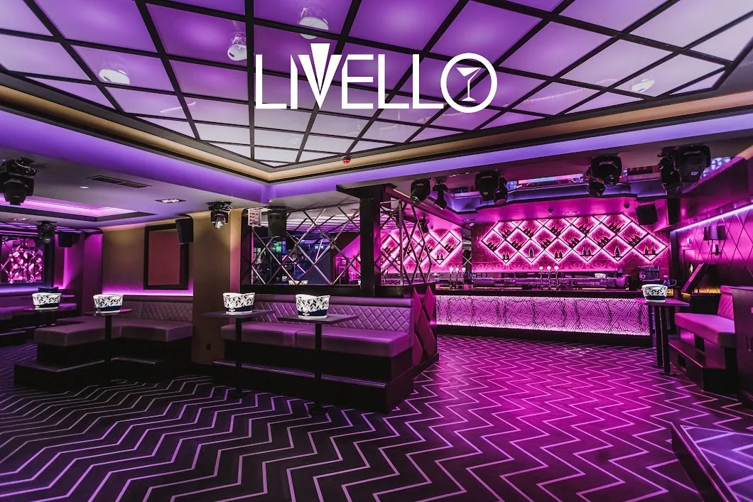 Interior of a modern nightclub with purple neon lighting, geometric ceiling design, velvet couches, illuminated tables, a bar area with patterned lighting, and a patterned floor.
