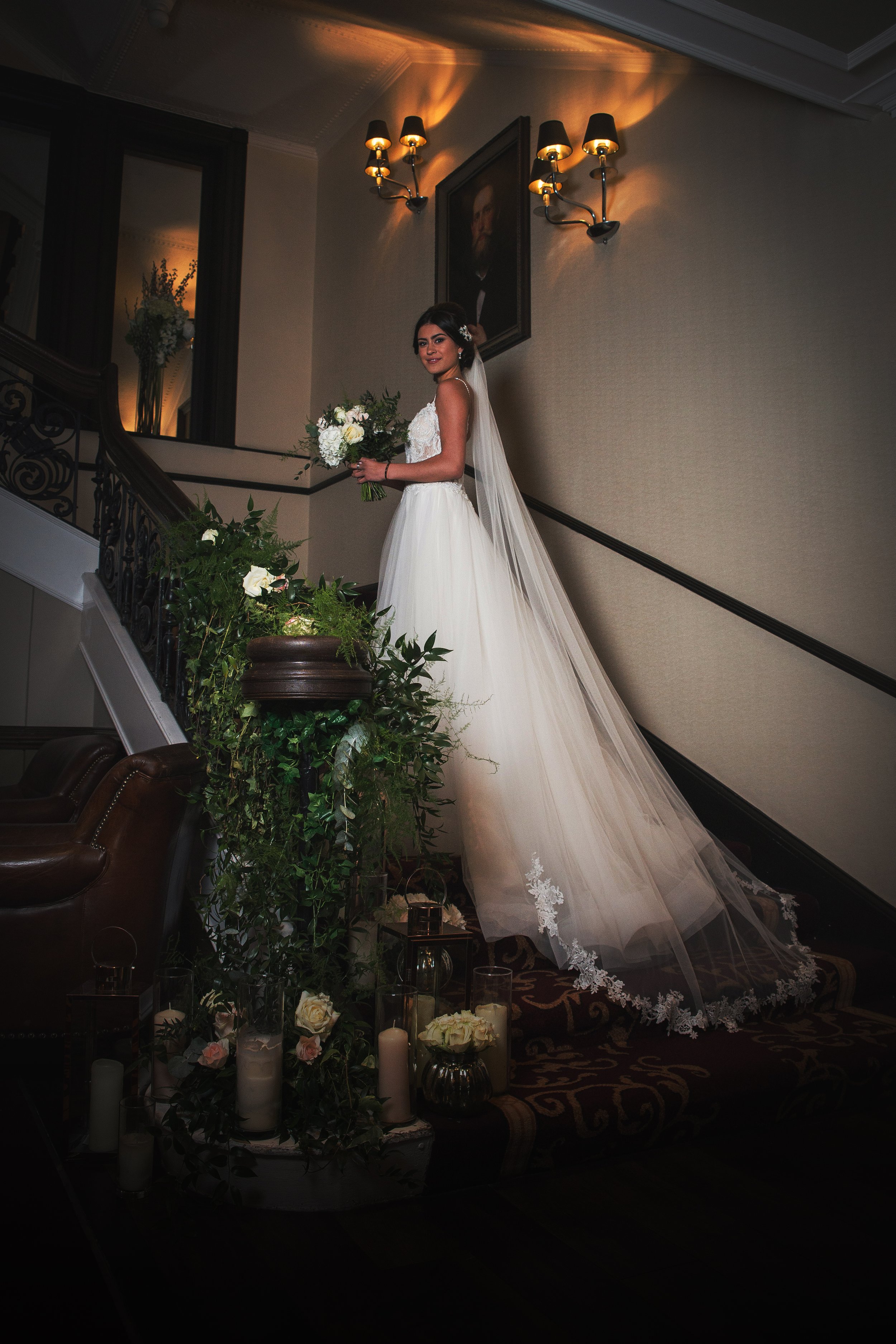 A bride in a white wedding dress holding a bouquet of white flowers stands on a staircase with decorative black railings. The staircase is adorned with candles and greenery at the bottom. The background features a framed portrait on the wall and elegant wall sconces with warm lighting.