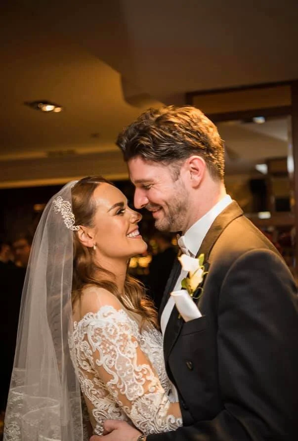 A bride and groom smiling and looking at each other during their wedding reception. The bride is wearing a lace wedding dress and veil, and the groom is in a tuxedo with a boutonniere.
