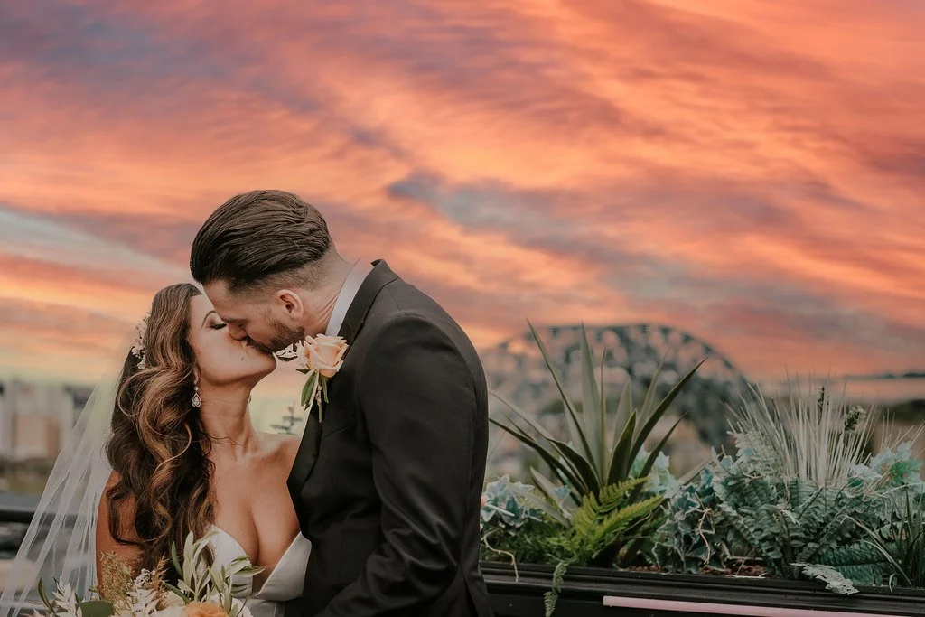A bride and groom kissing at sunset with a colorful sky in the background and plants in the foreground.