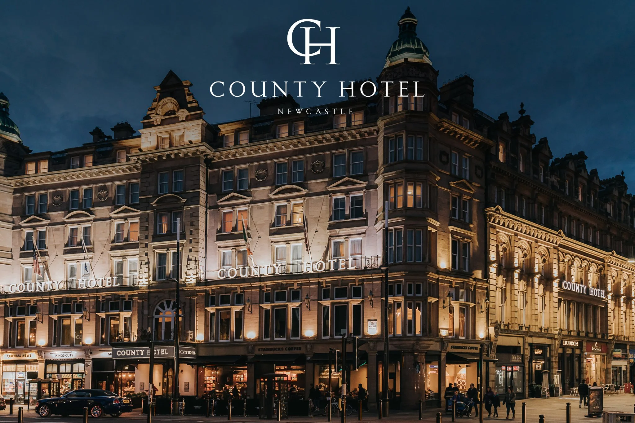 Night view of the historic County Hotel building in Newcastle illuminated with warm lighting, featuring multiple ornate rooftops and decorative facades, with people walking and cars parked in front.