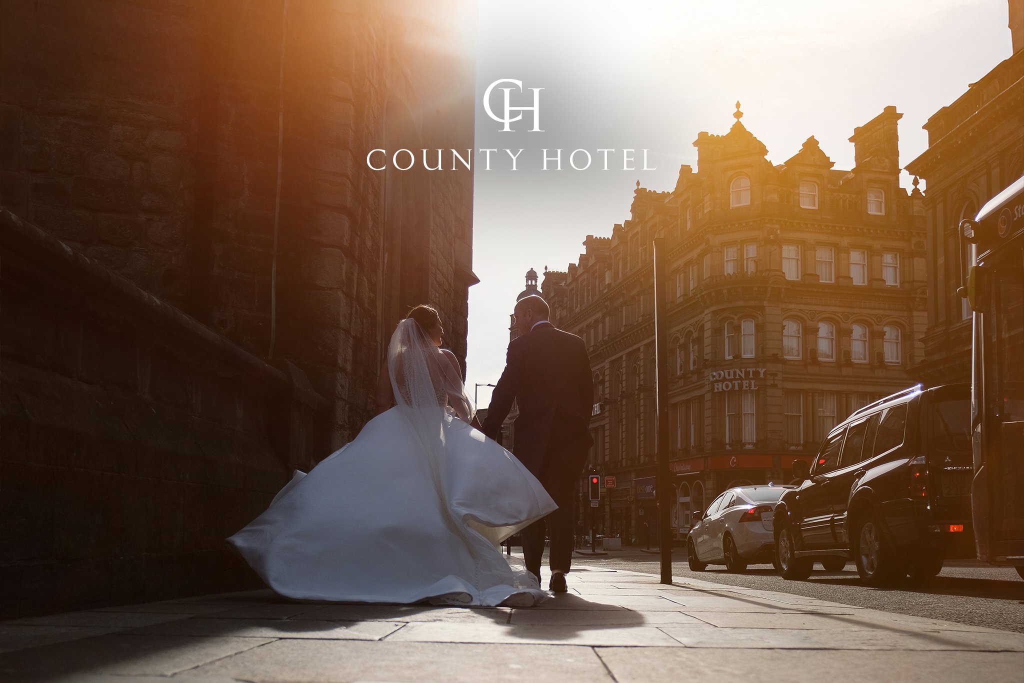 A bride and groom holding hands and walking in a city street during sunset, with a large building labeled 'County Hotel' and parked cars along the street.