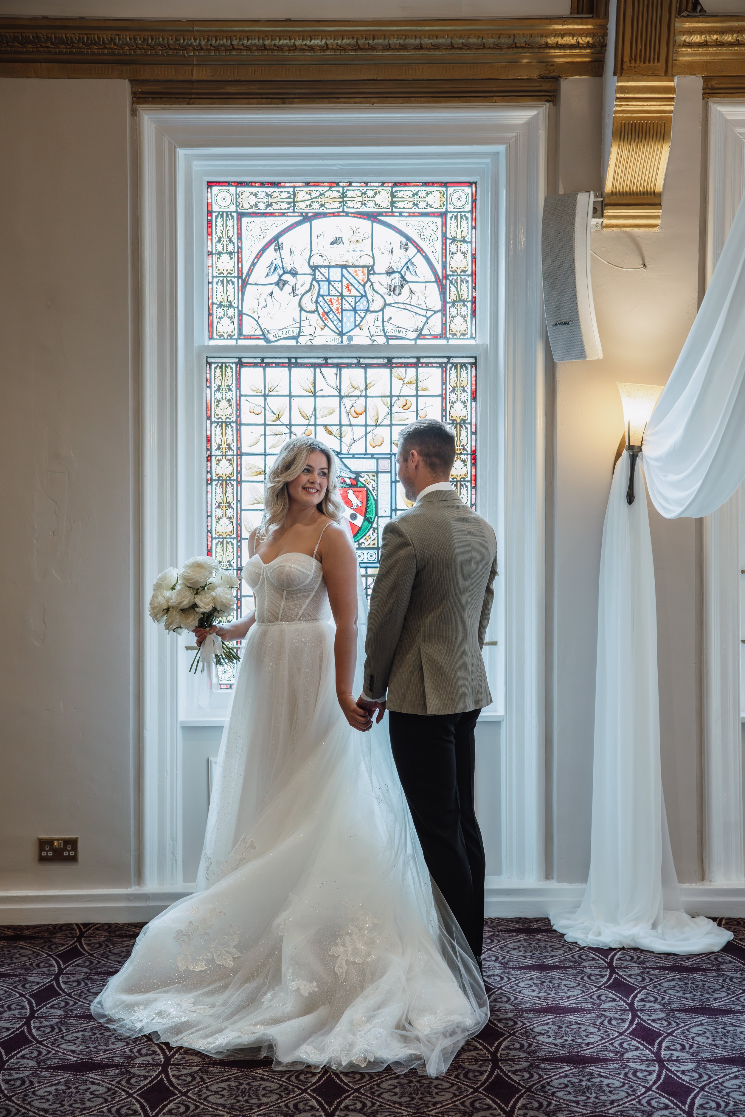 A bride and groom holding hands in a room with stained glass windows. The bride is wearing a white wedding gown and holding a bouquet of white flowers, while the groom is dressed in a gray suit. They are smiling at each other.