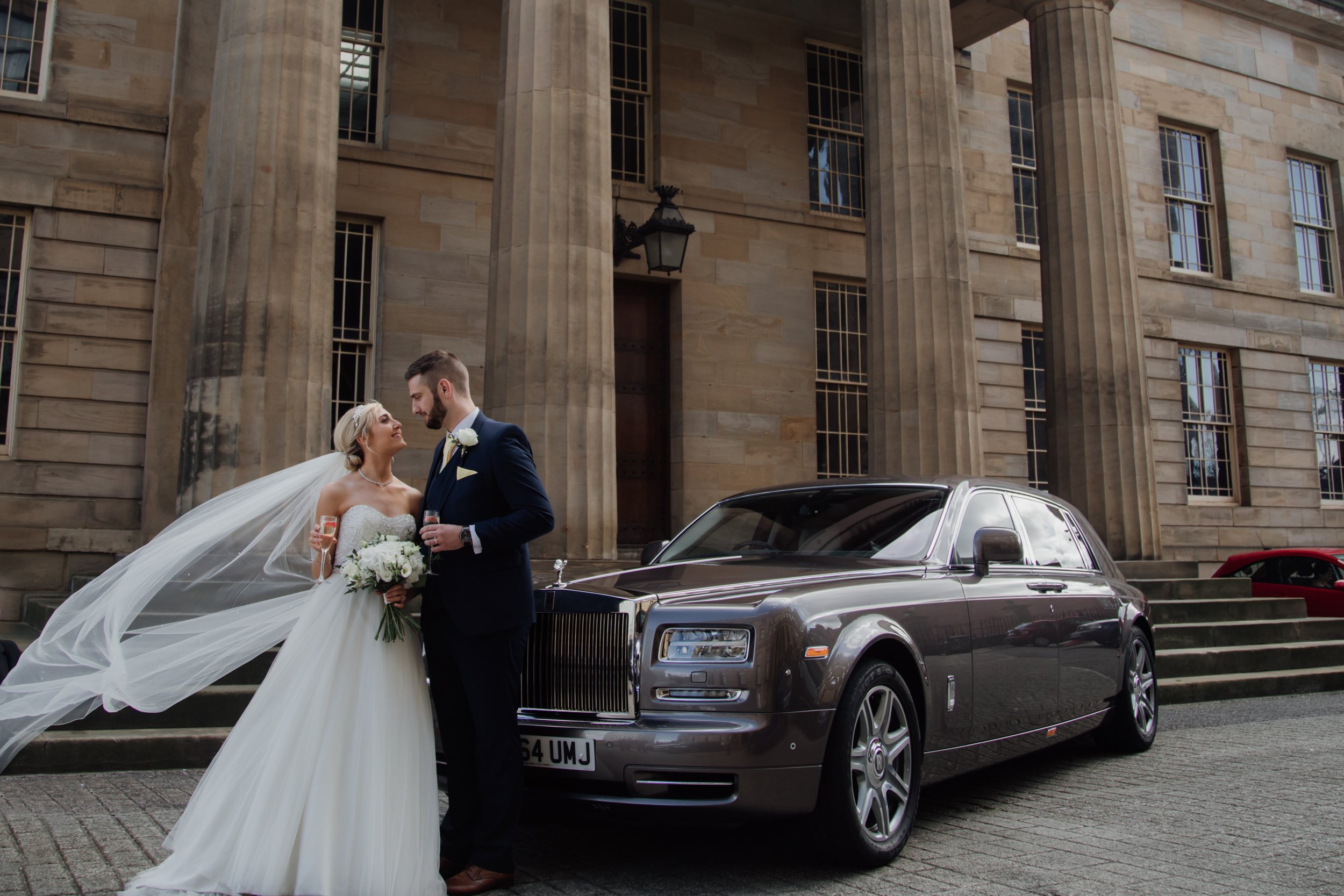 Bride and groom standing in front of a vintage car outside a large stone building, with the bride holding a bouquet and wearing a long wedding gown and veil, while the groom is dressed in a dark suit with a boutonniere.