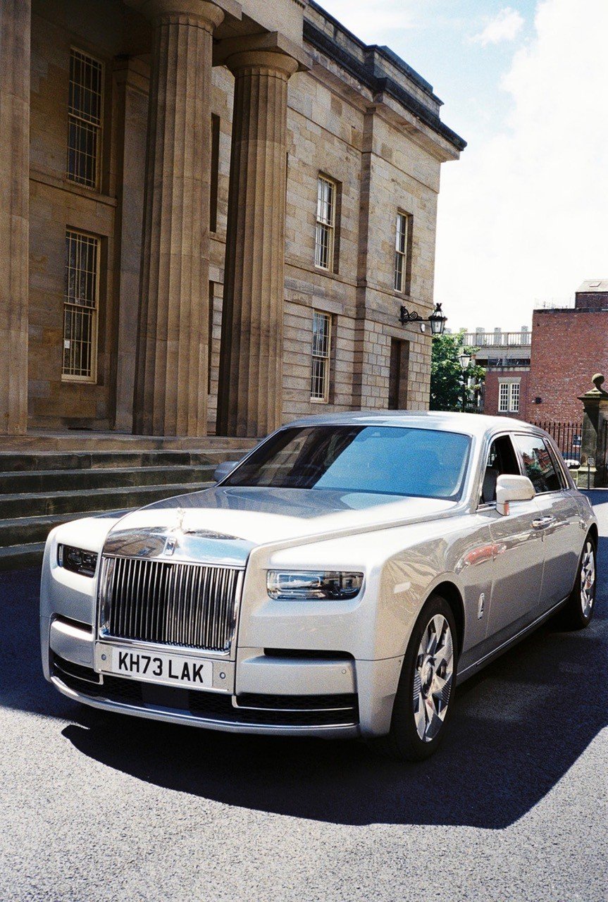 A white luxury Rolls-Royce car parked on a city street in front of a historic building with large columns and tall windows.