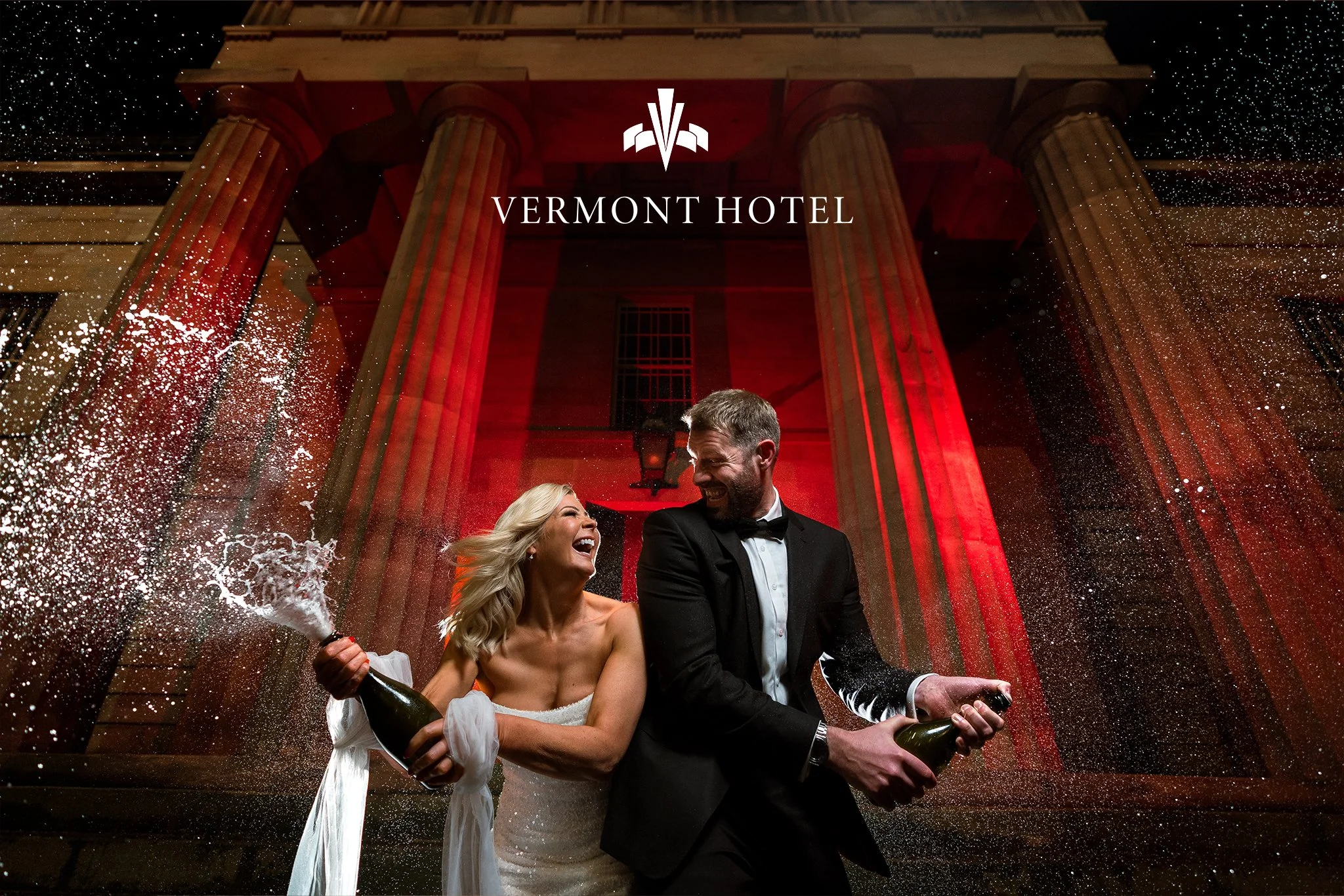 A couple in wedding attire celebrating with champagne on the stairs of the Vermont Hotel at night, with water splashing around them and a large brick building illuminated by red lights.