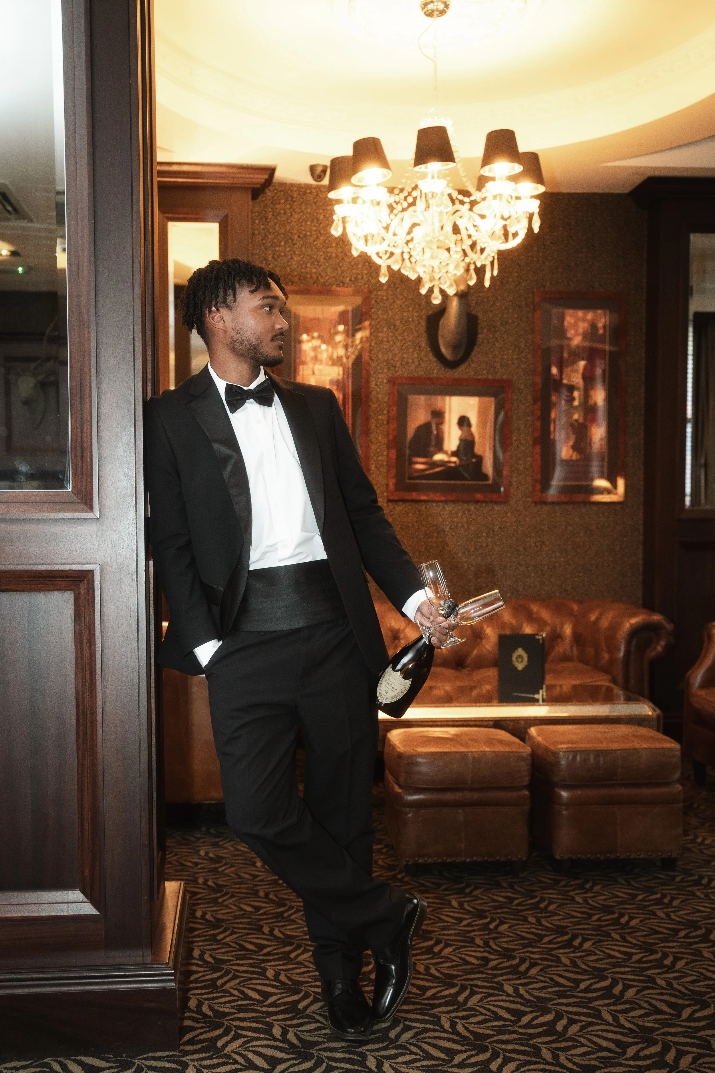 A man in a tuxedo holding a bottle of champagne and a glass in a luxurious, dimly lit room with ornate decor, a chandelier, and framed pictures on the wall.