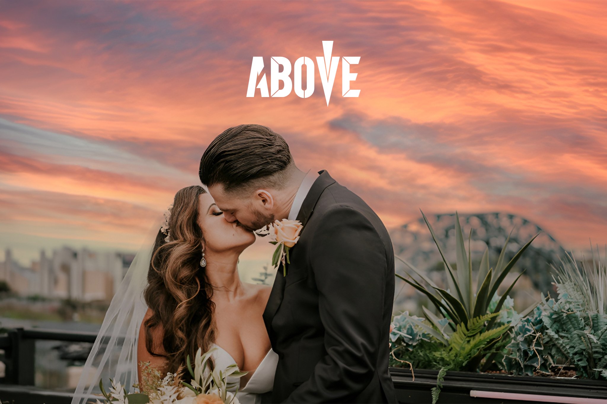 A couple, dressed in wedding attire, sharing a kiss during sunset on a rooftop with city buildings in the background.