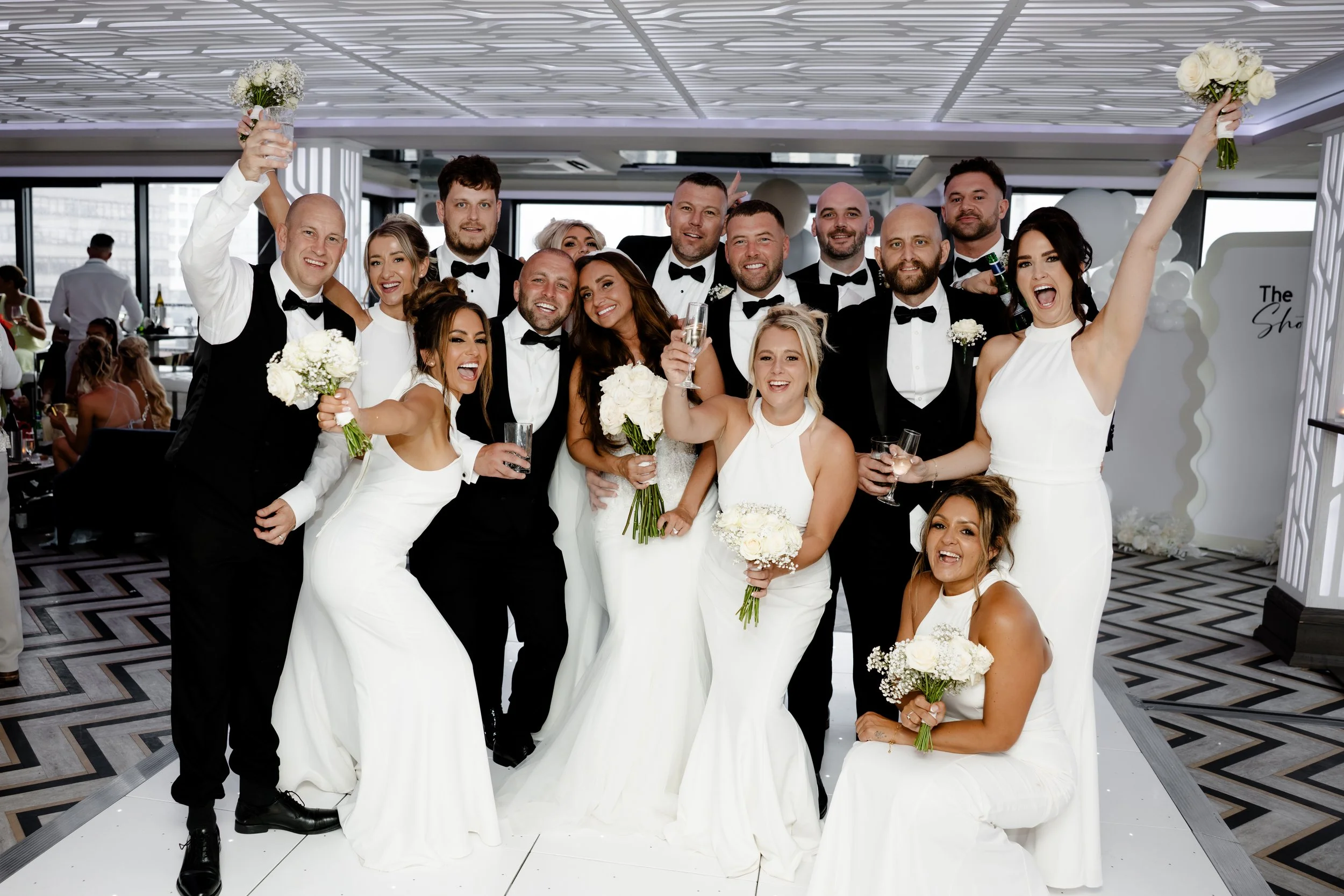 A group of people at a wedding celebration, dressed in formal attire, smiling and holding bouquets and drinks, indoors with a modern decorated background.