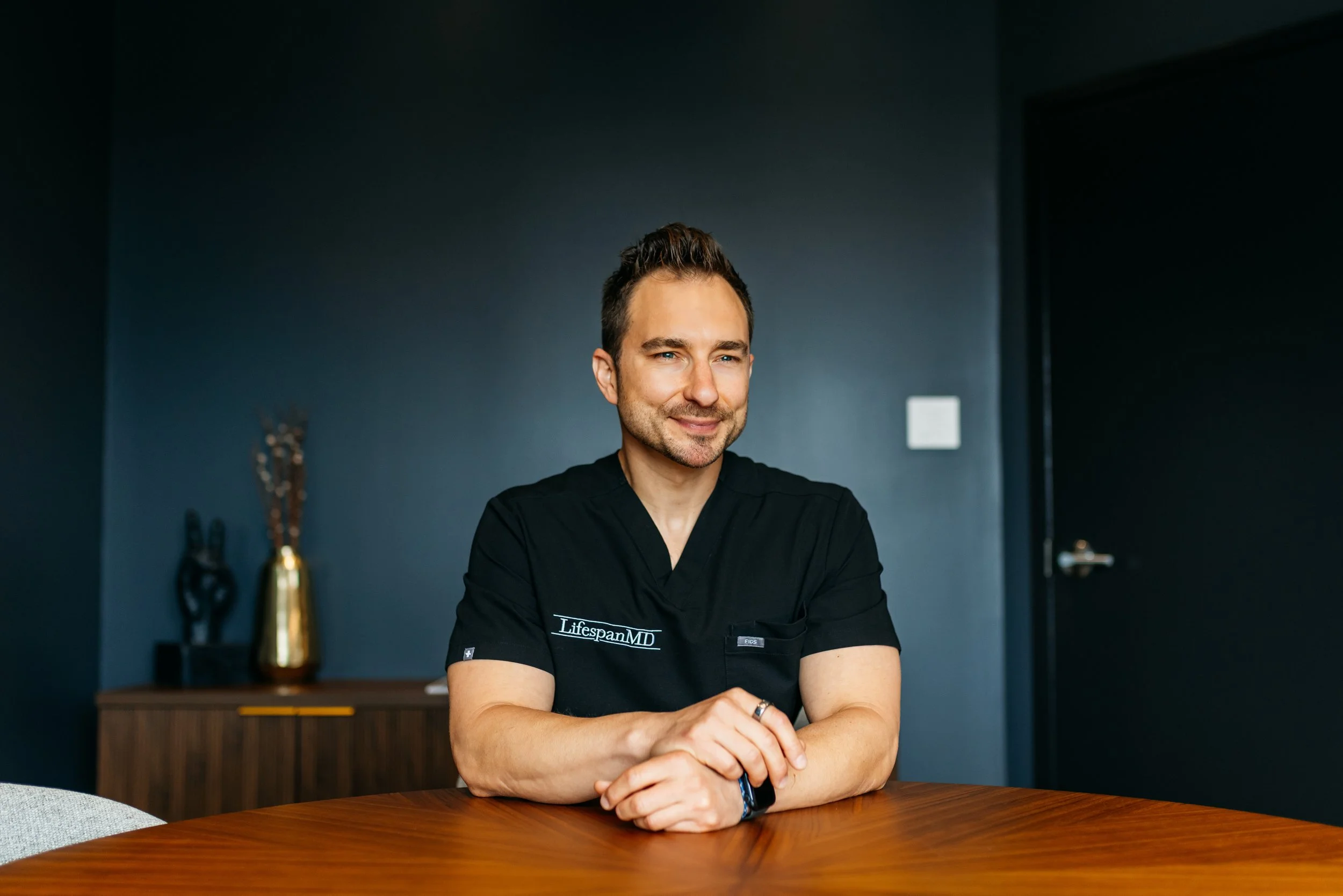 A man with dark hair and blue eyes sitting at a wooden table, wearing a black medical uniform with 'Lifespan MD' on it, smiling at the camera in a room with dark blue walls.