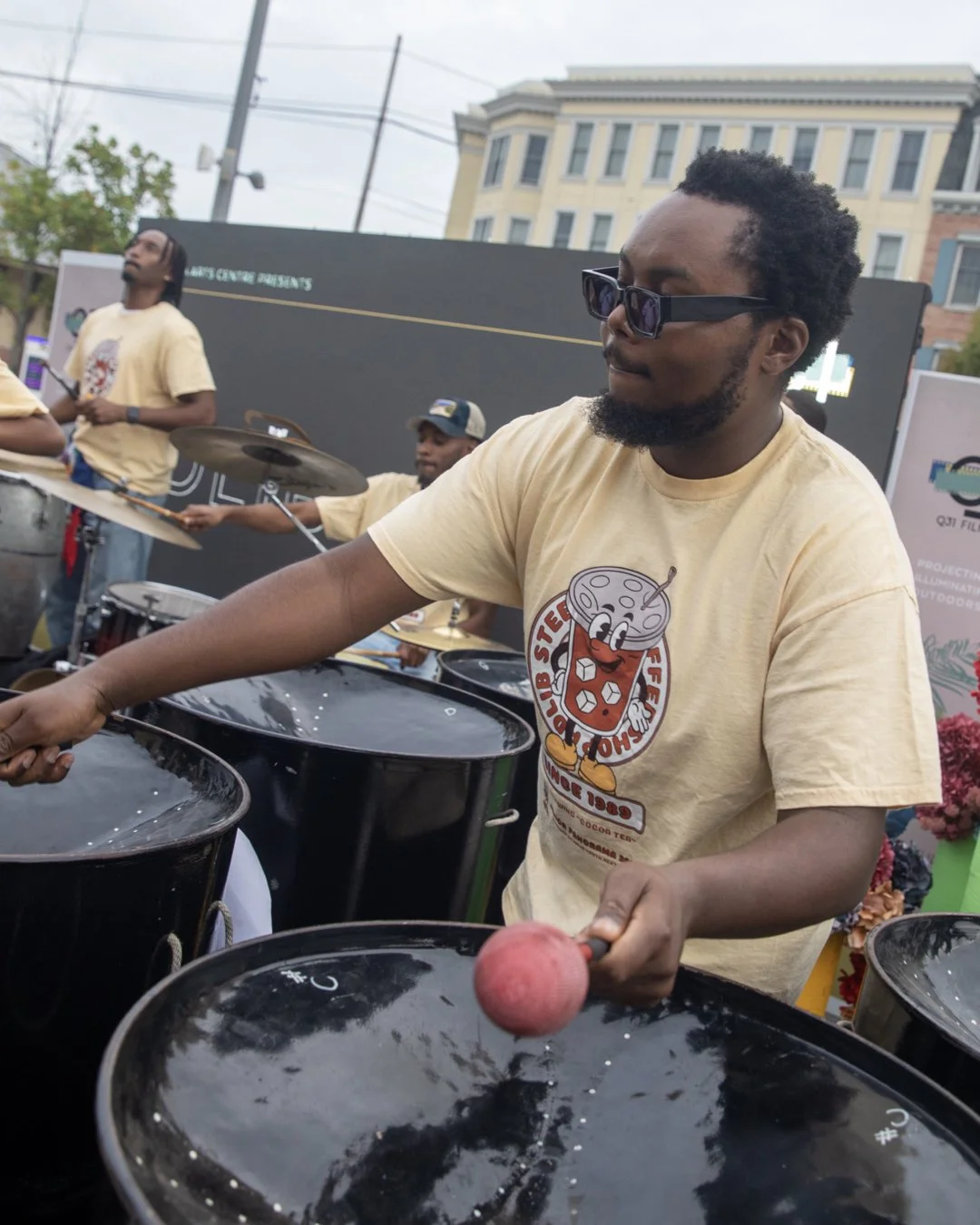 A man wearing sunglasses and a beige T-shirt playing drums outdoors at a street event, with other musicians and a building in the background.