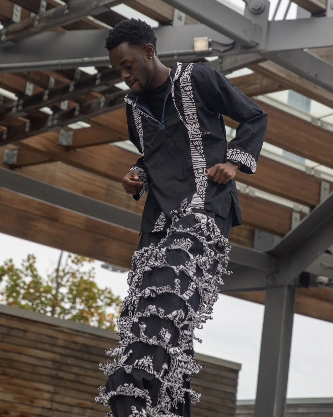 A young man with black curly hair is standing outdoors under a wooden pergola, looking down in a thoughtful pose. He is wearing a black shirt and black pants with white patterned ruffles, and appears to be deep in thought or contemplation.