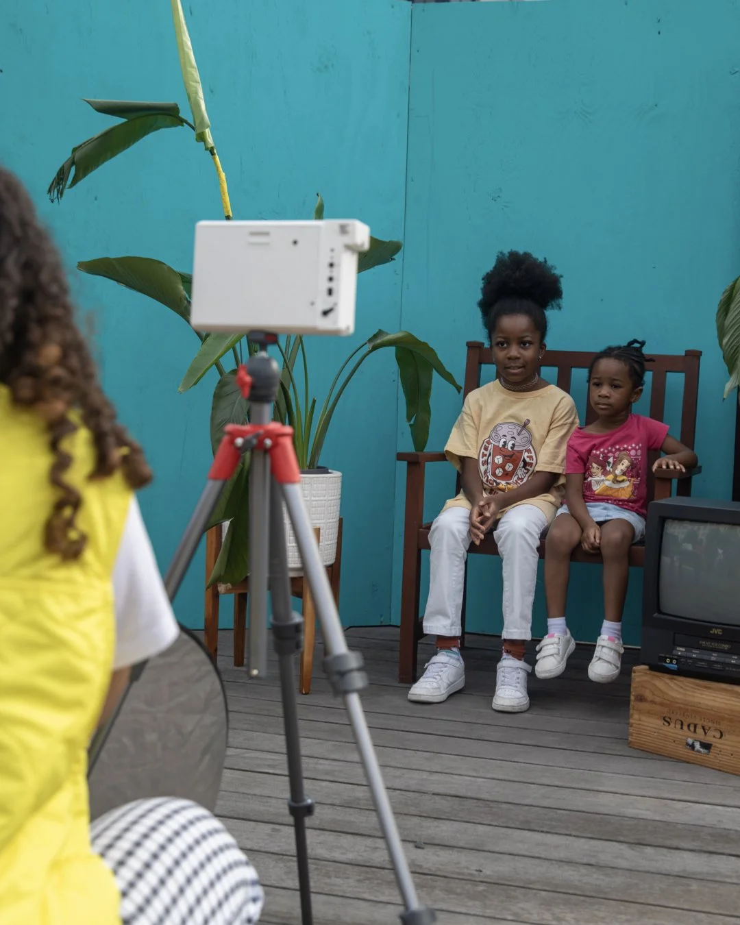 Two young girls sitting on a wooden bench in a room with teal walls and large leafy plants, while a person with braided hair wearing a yellow shirt takes a photo or video with a camera mounted on a tripod.