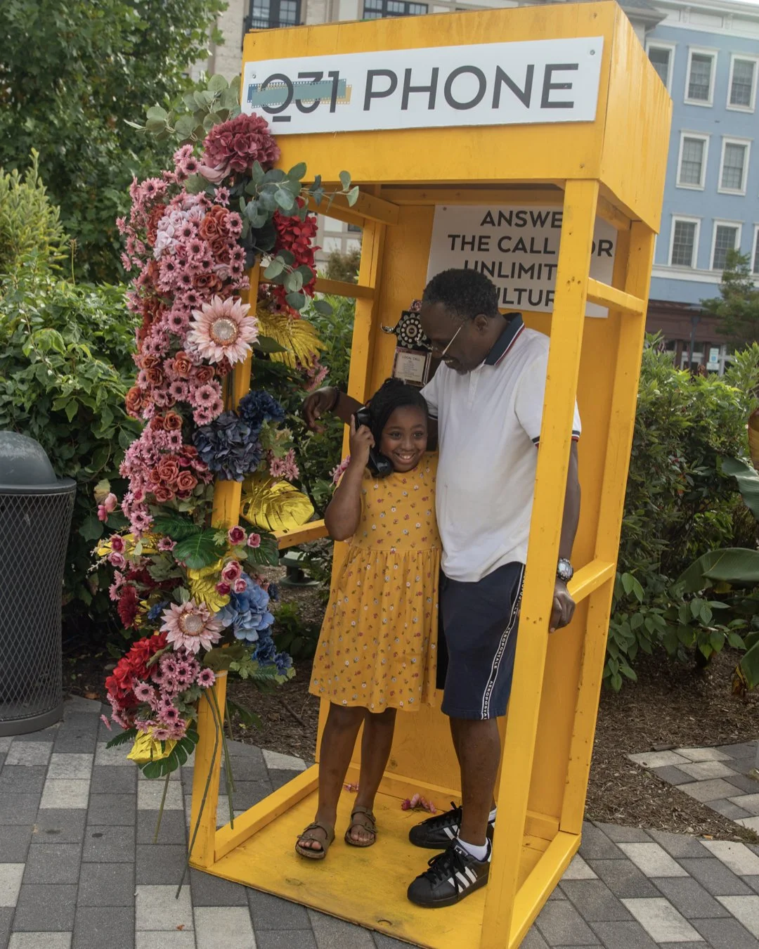 A man and girl standing in a yellow phone booth decorated with pink, red, and blue flowers, smiling and talking on the phone.