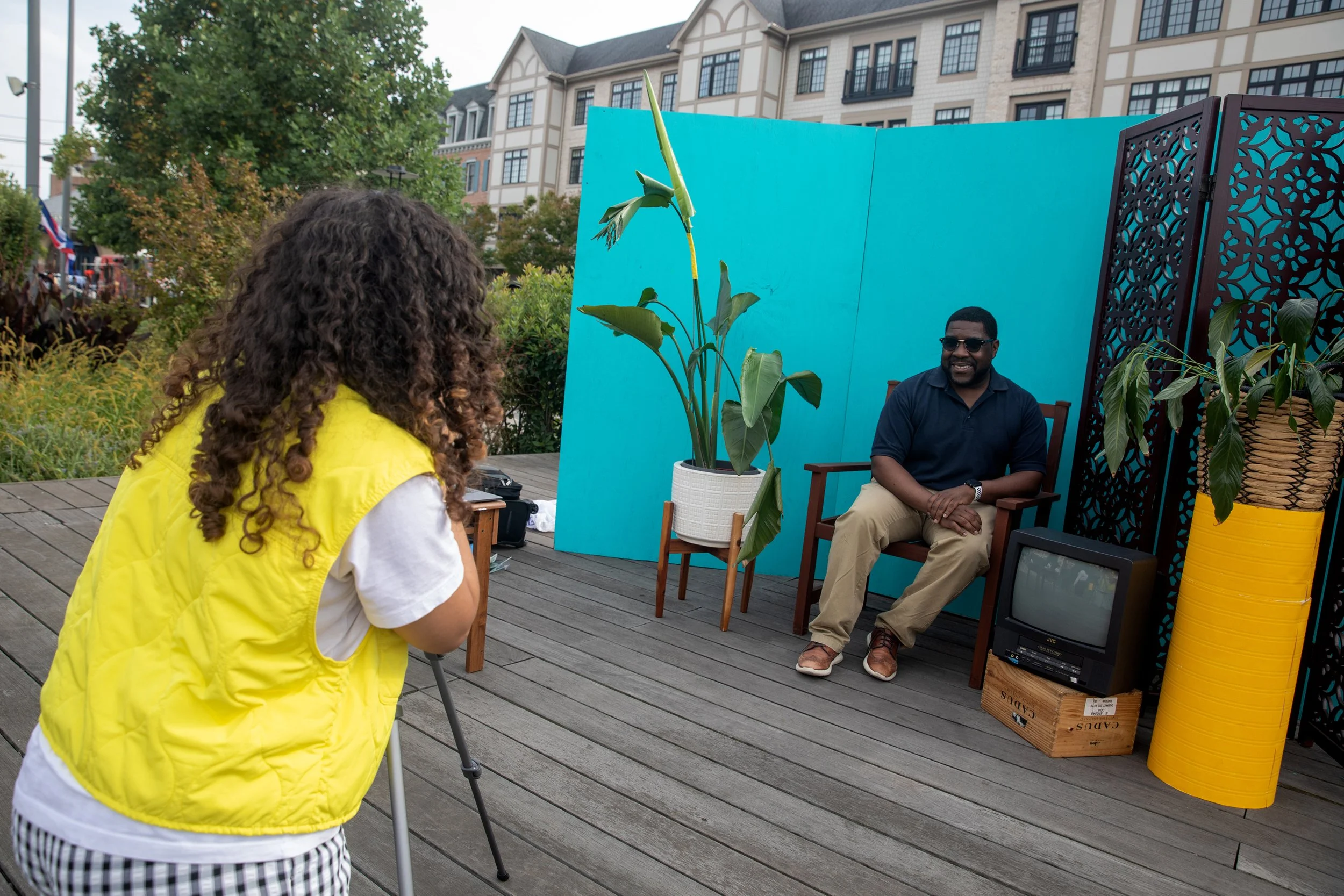 A woman with curly hair wearing a yellow vest and striped pants taking a photo or video of a man sitting on a chair behind a decorative divider at an outdoor event in a residential area with apartment buildings and trees in the background.