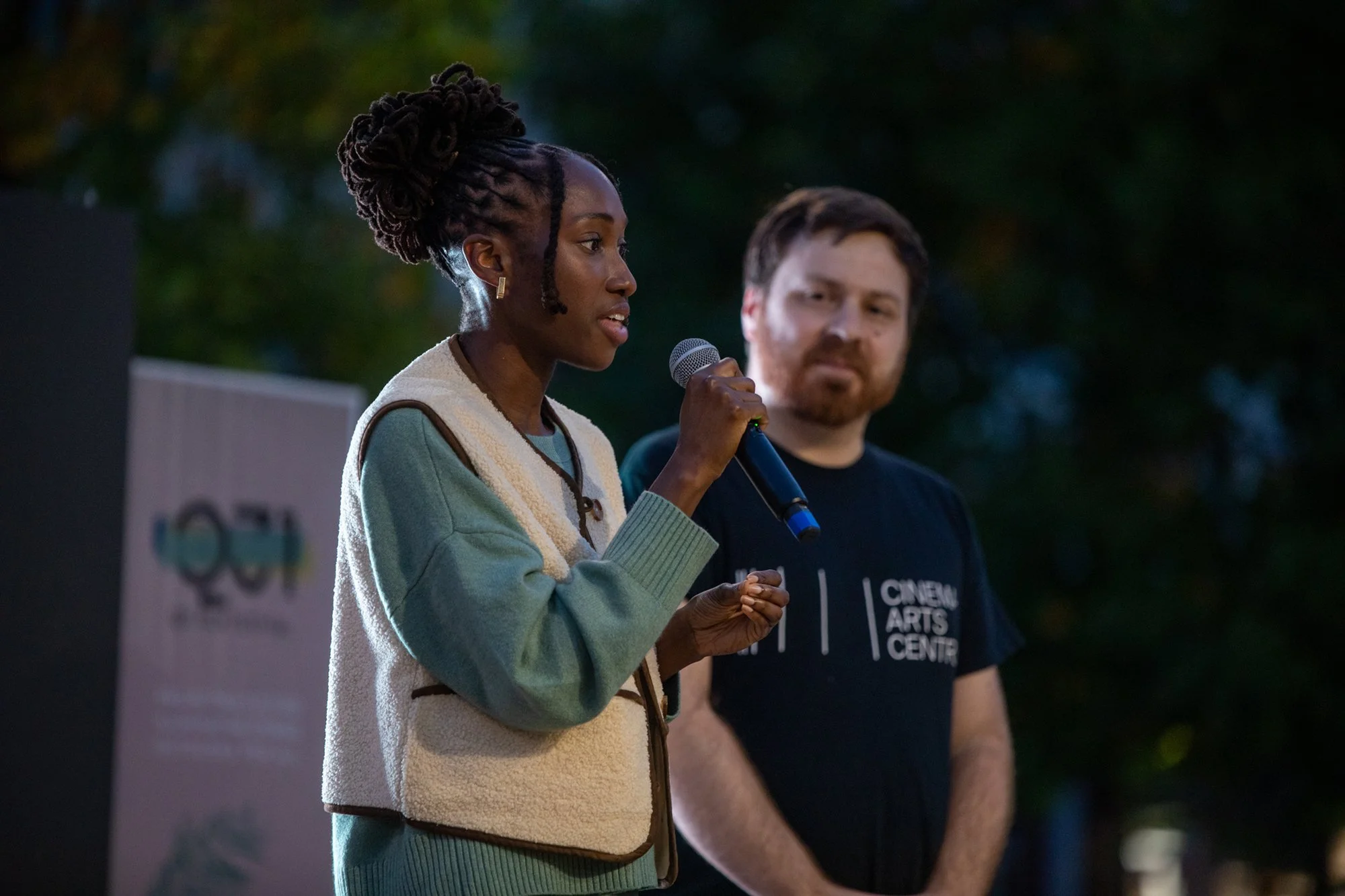 A woman with dark skin, braided hair styled in a bun, wearing earrings and a cozy jacket, is speaking into a microphone during an outdoor event at dusk, with a man standing beside her.