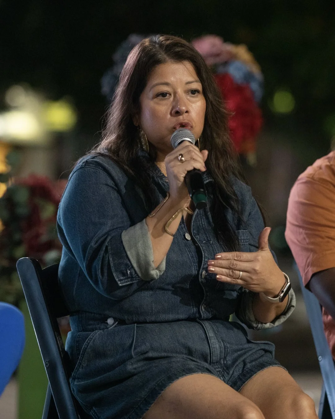 A woman with long dark hair wearing a denim jacket and shorts, holding a microphone and speaking at an outdoor event at night, with blurred colorful flowers in the background.