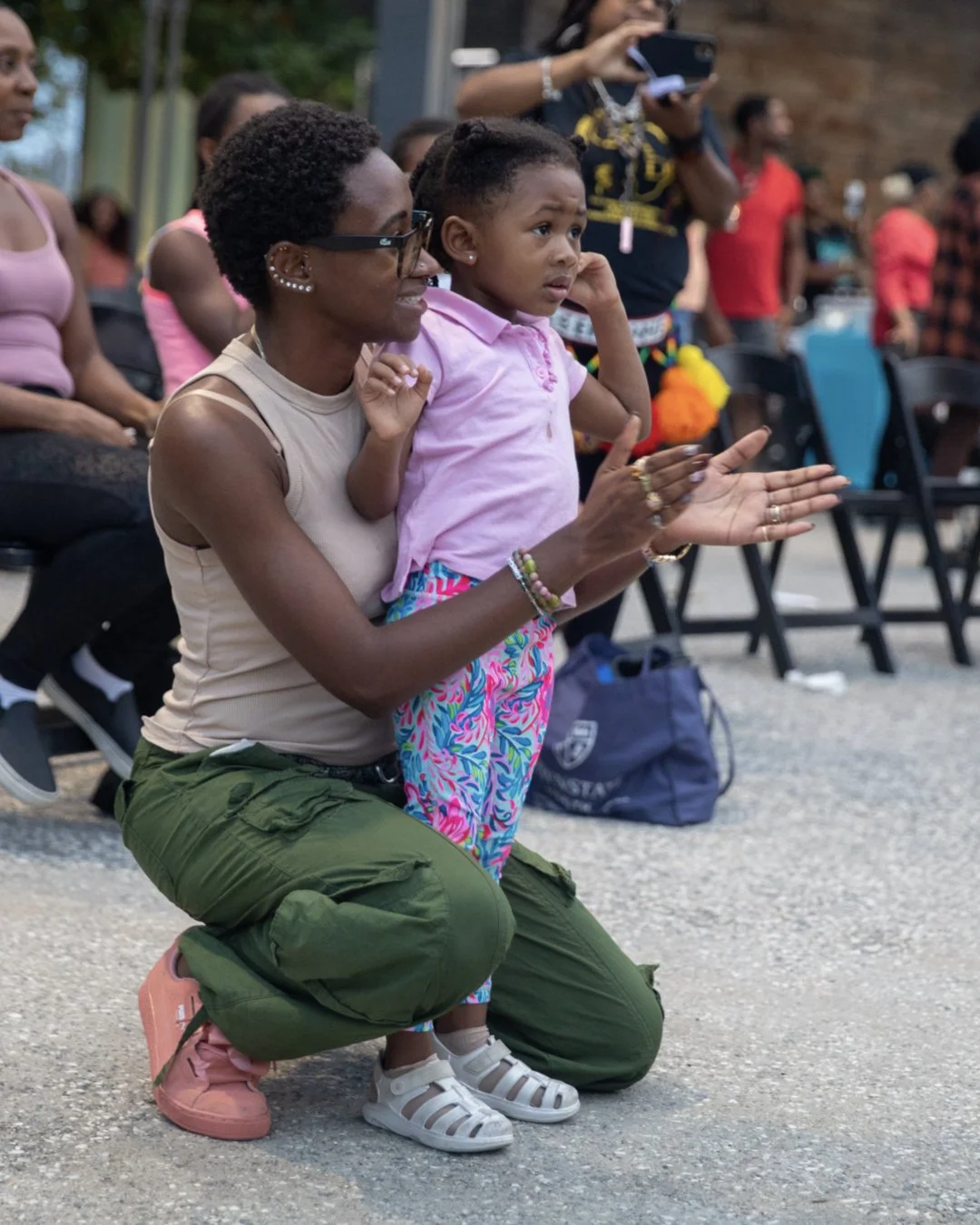 A woman kneeling on the ground with a young girl standing beside her, both looking attentively at something in front of them. The woman is smiling, wearing a beige sleeveless top, green cargo pants, and pink shoes. The girl looks curious, touching he