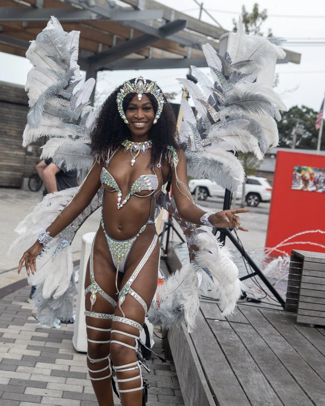 Woman in elaborate feathered carnival costume with sparkling accessories, smiling at the camera, outdoors on a paved area with cars and a red backdrop in the background.