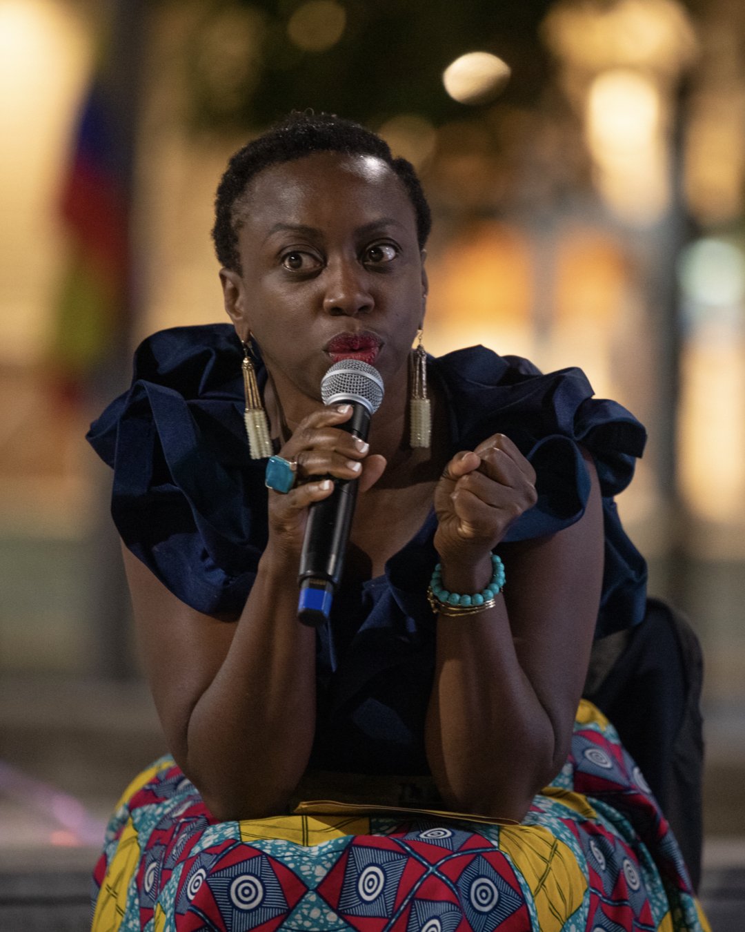 A woman with short hair and wearing a dark blue dress with puffed sleeves is holding a microphone and speaking. She is wearing colorful jewelry and sitting in a space with blurred warm lighting in the background.