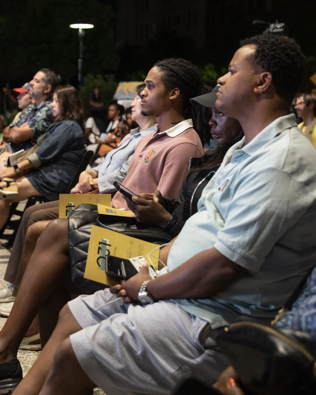 Audience seated outdoors at night, attentively watching the films, some holding informational brochures and using their phones.