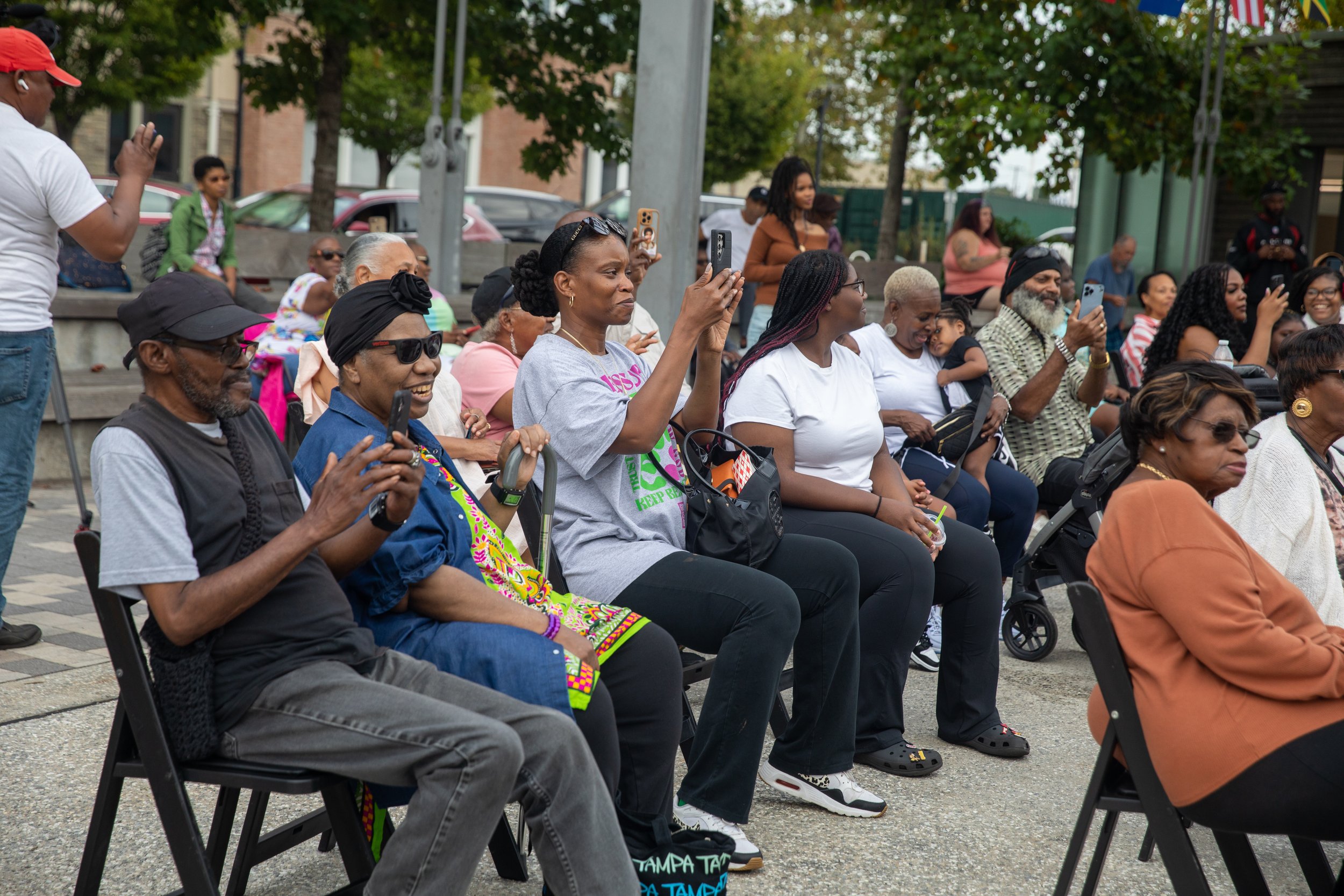 Group of diverse people sitting and standing in outdoor public space, some taking photos or videos with their phones during a community event.