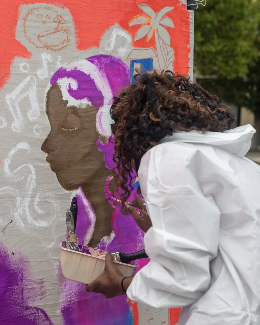 A person with curly hair wearing a white jacket paints a mural of a woman's silhouette with purple and pink hair, singing with music notes around her on a wall outdoors.