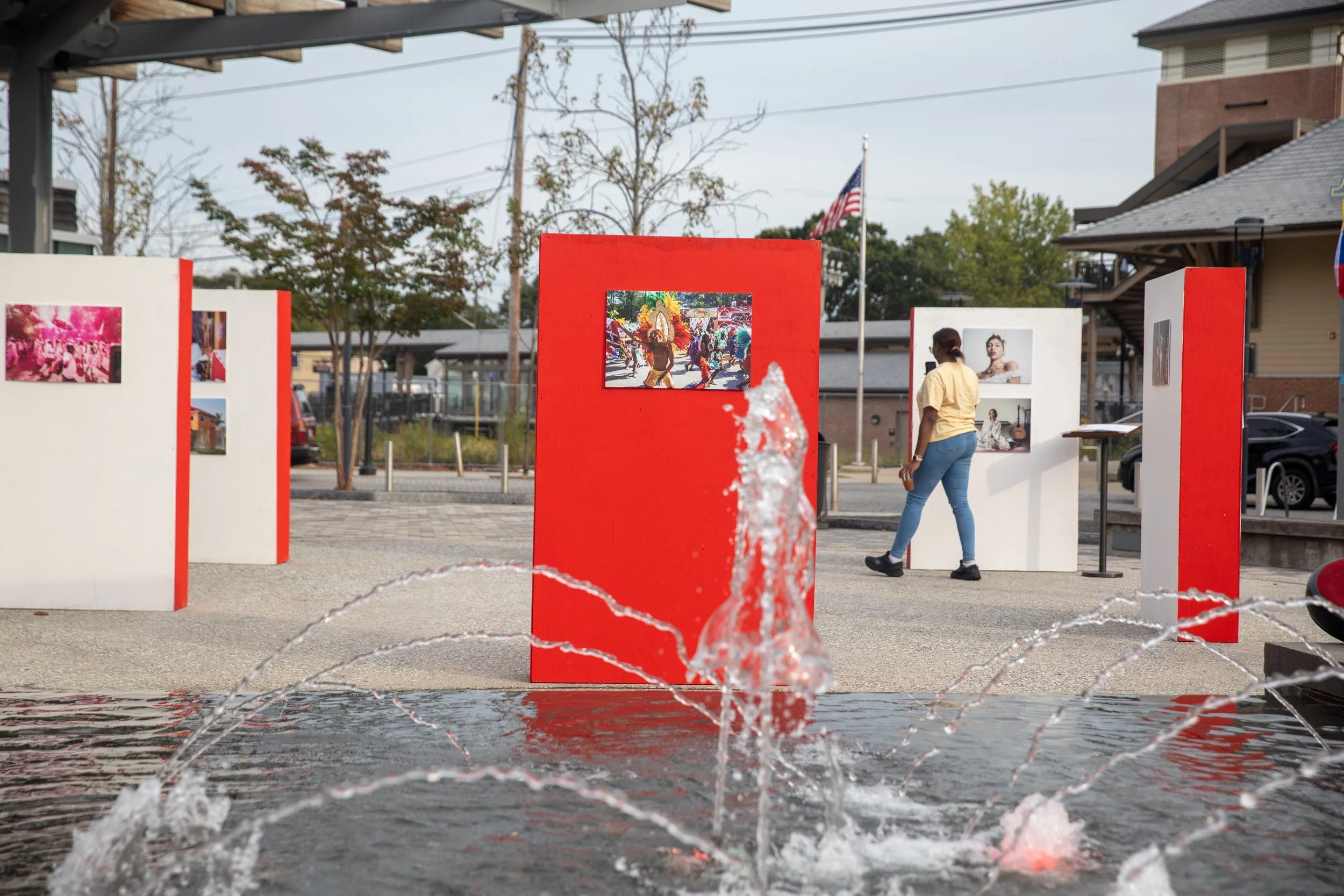 A fountain with water splashing in front of outdoor exhibition panels, including one showing a colorful parade, with a woman walking nearby and a neighborhood background with trees, parking, and a flagpole.