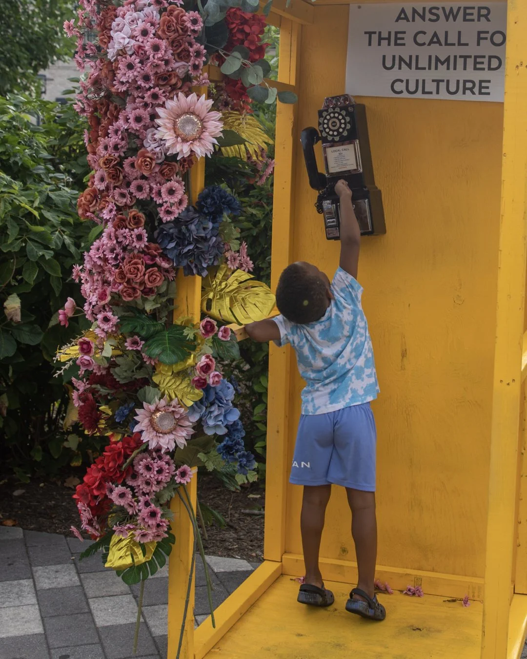 A young boy in a blue and white tie-dye t-shirt and blue shorts is dialing a payphone mounted on a bright yellow phone booth. The booth is decorated with a large, colorful floral arrangement featuring pink, red, purple, and blue flowers and green leaves. A sign on the booth reads, "ANSWER THE CALL FOR UNLIMITED CULTURE."