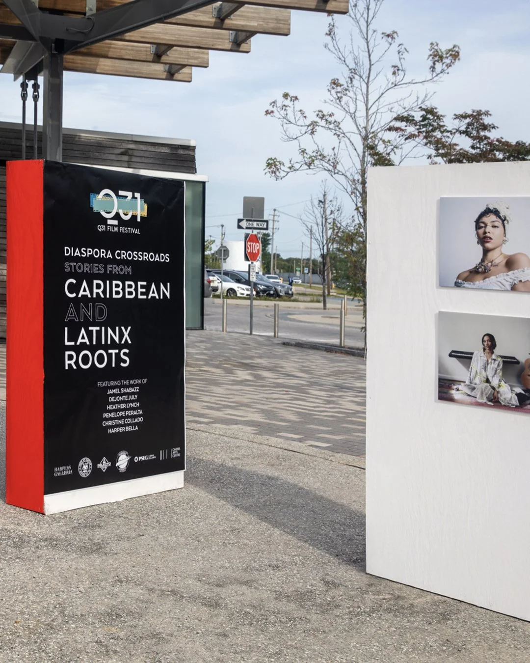A billboard advertising the Q3 Film Festival, highlighting stories from Caribbean and Latinx roots, with features of various artists and cultural references. Next to it, there are photographs of women on a white display panel outdoors near a parking 