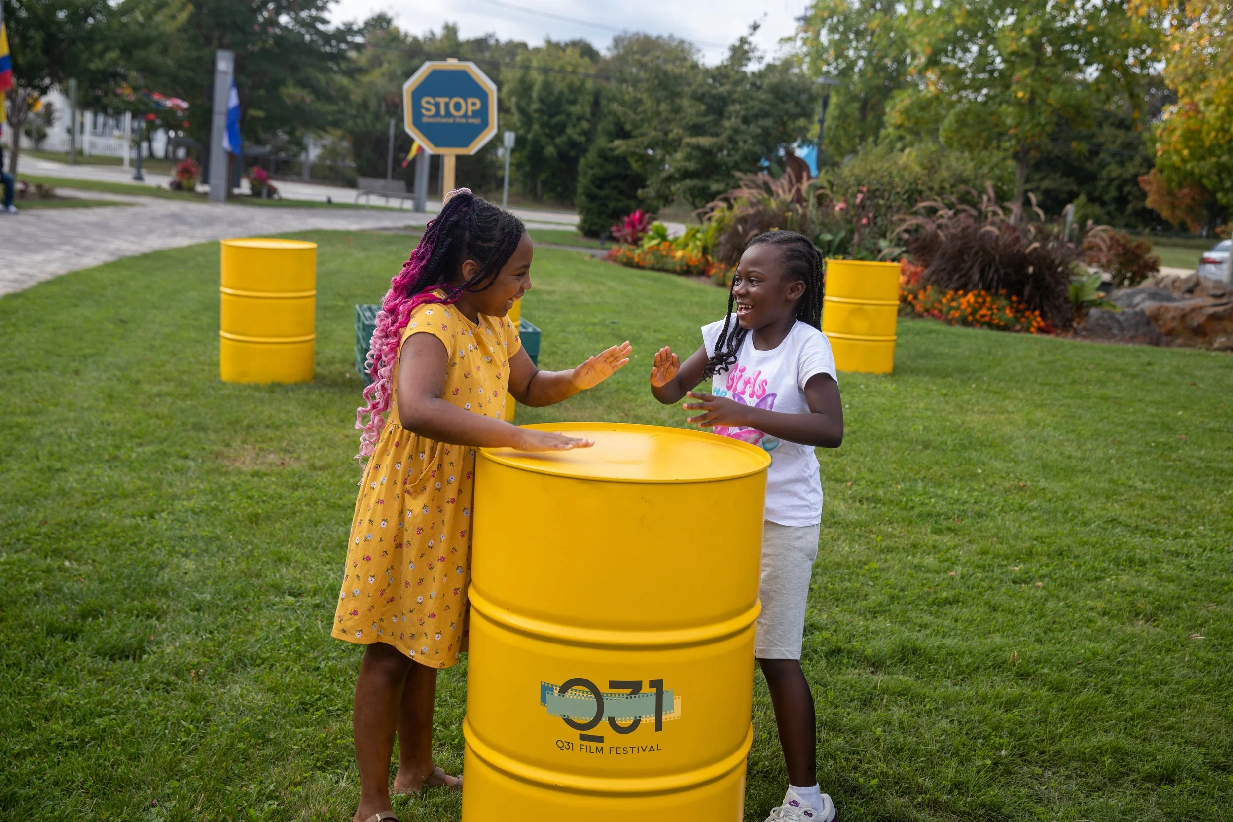 Two young girls with braided hair smiling and high-fiving each other, standing behind a yellow barrel with the Q31 Film Festival logo, outdoors on green grass with trees and a sidewalk in the background.