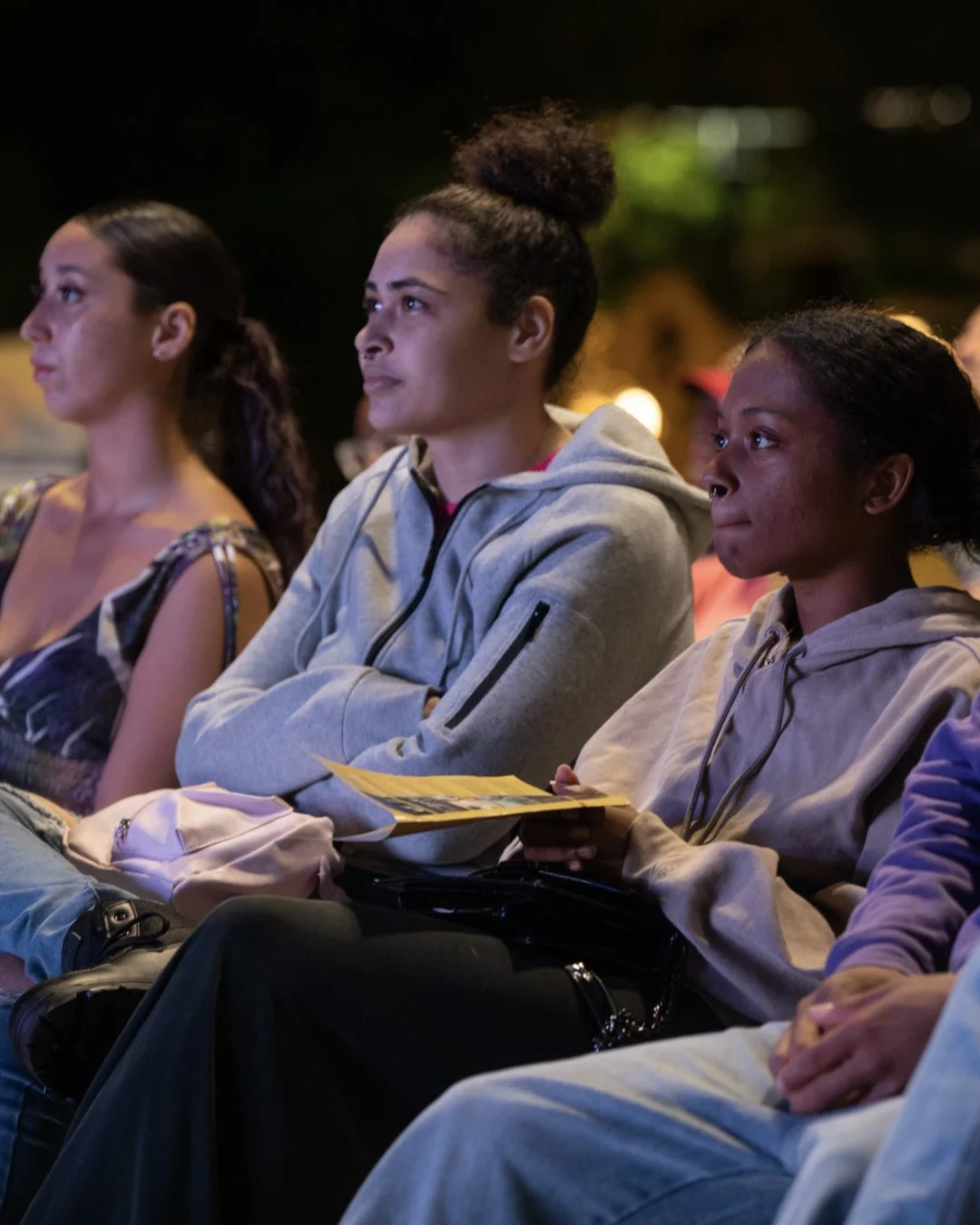 Three women sitting outdoors at night, attentively watching an event, with a woman in the middle holding a yellow pamphlet or brochure.