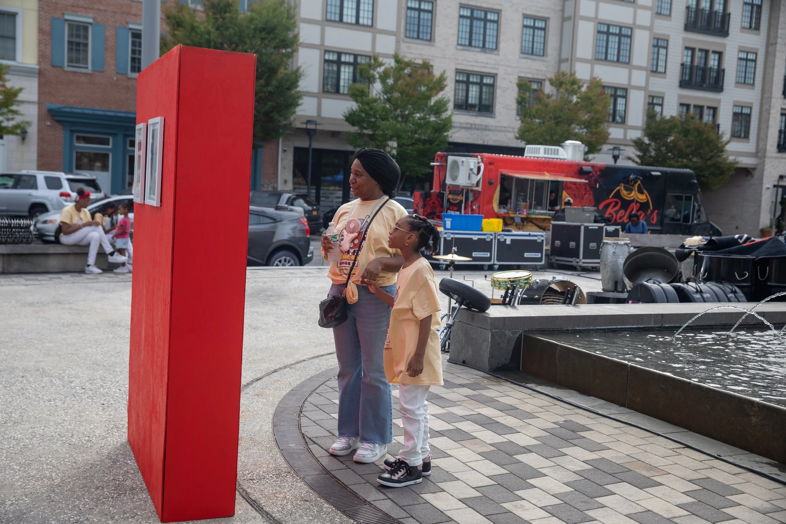A woman and a girl stand next to a bright red rectangular art installation in an urban park, with a fountain in the foreground and a food truck in the background. The woman holds a water bottle and wears a beige t-shirt with a design, while the girl wears a gold-colored top and white pants. There are trees, parked cars, and a building in the background.