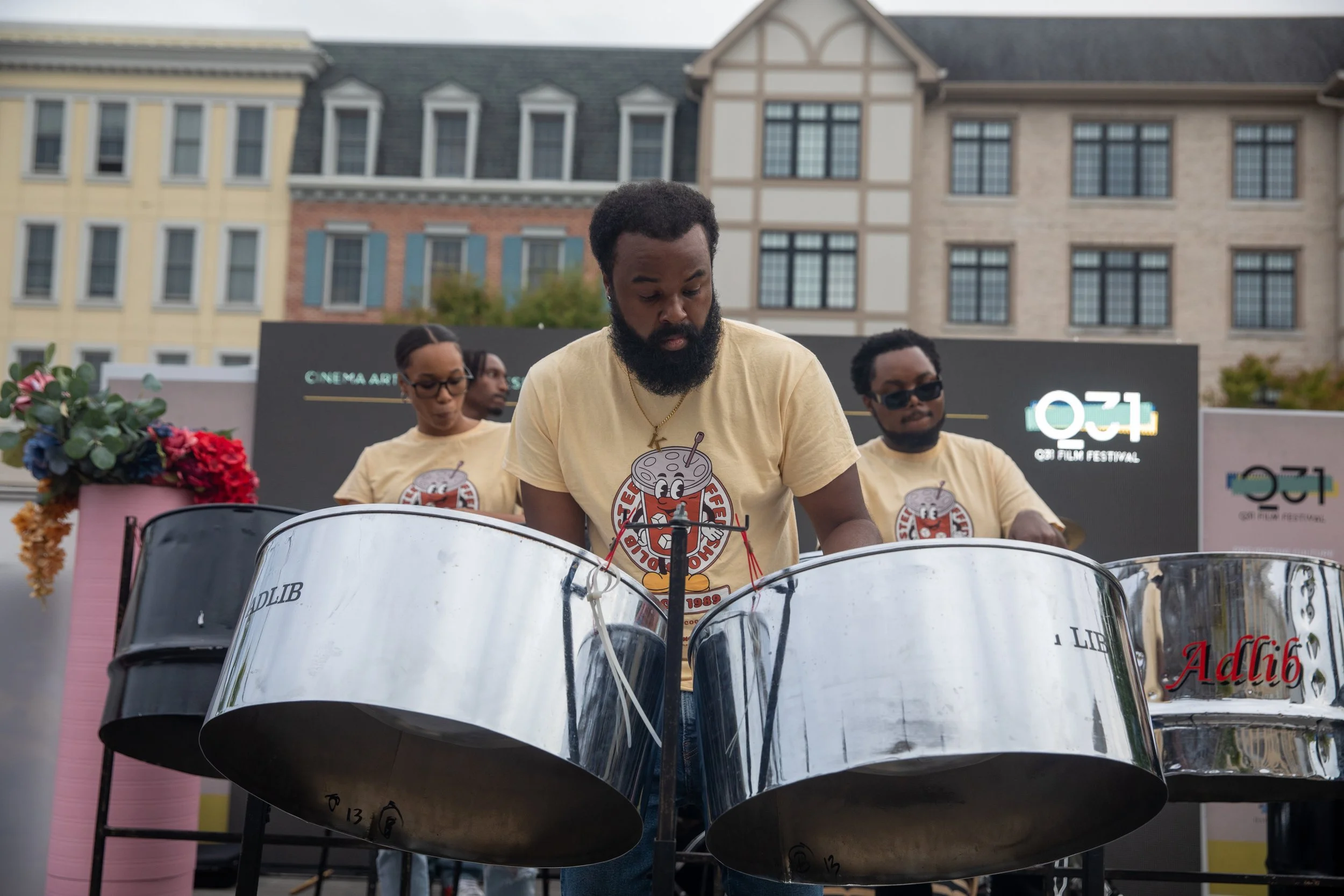 Three festival musicians playing steel drums outdoors in front of multi-story buildings, wearing matching beige t-shirts with a cartoon graphic and text, with a sign for the O'z Film Festival in the background.