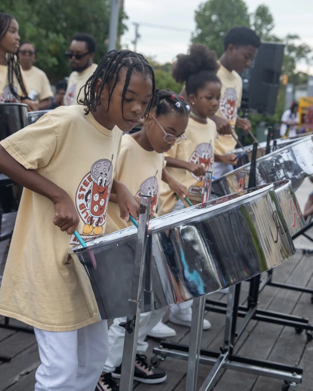 Children playing steel drums at an outdoor event, all wearing matching beige t-shirts with a graphic design.