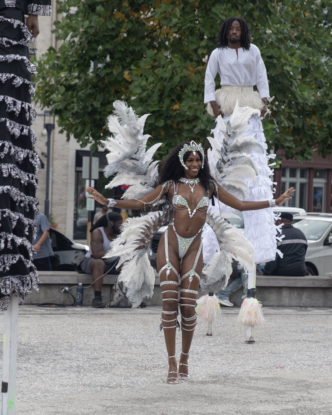 A woman in an elaborate carnival costume with feathers and sparkling jewelry, smiling and performing in a street parade, with a man on stilts dressed in white in the background.