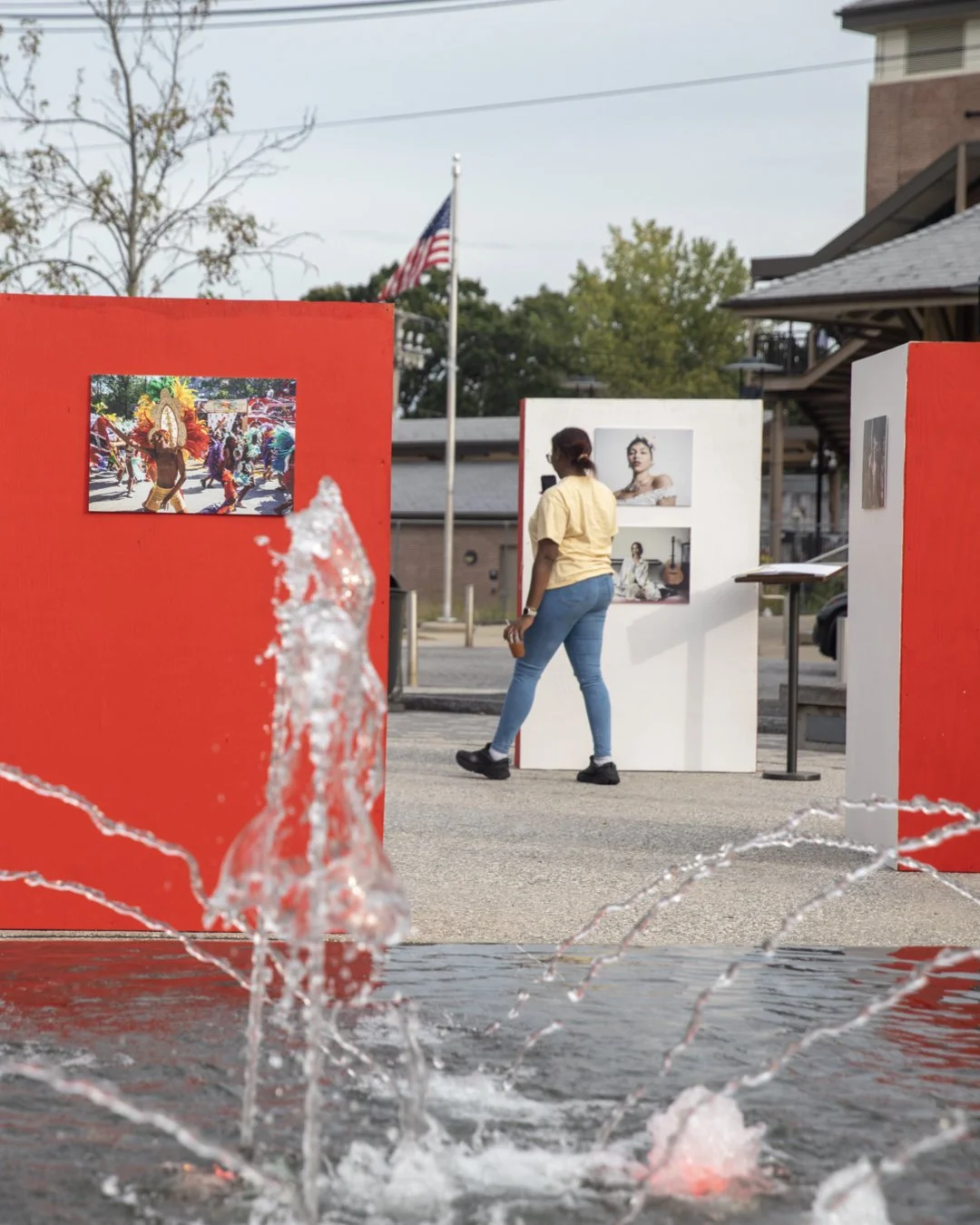 An outdoor art exhibition with photographs displayed on red and white panels, and a woman viewing the artwork. A water fountain in the foreground splashes water, with a background of trees, buildings, and an American flag.