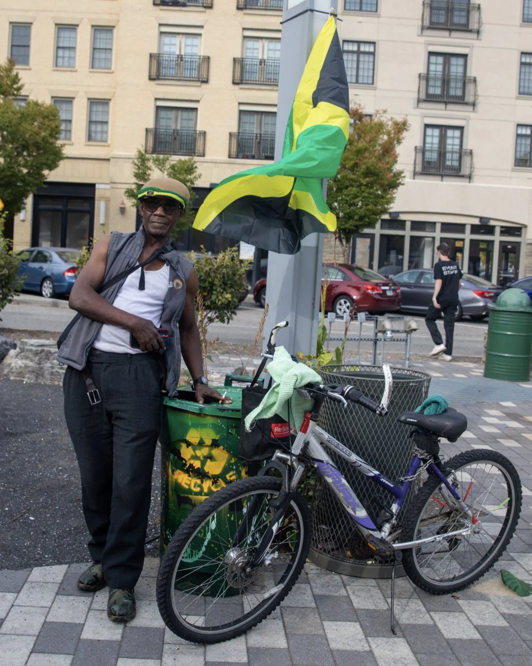 An older man in casual clothes and sunglasses standing beside a bicycle and a green trash bin with a flag attached, on a city sidewalk with buildings and parked cars in the background.