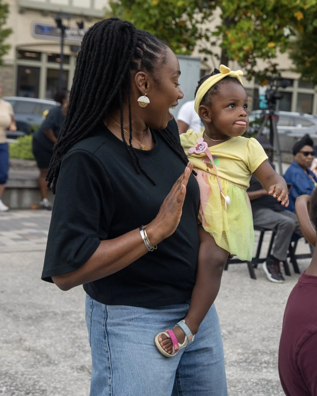 A woman with long dreadlocks holding a young girl in a yellow dress and headband at an outdoor event.